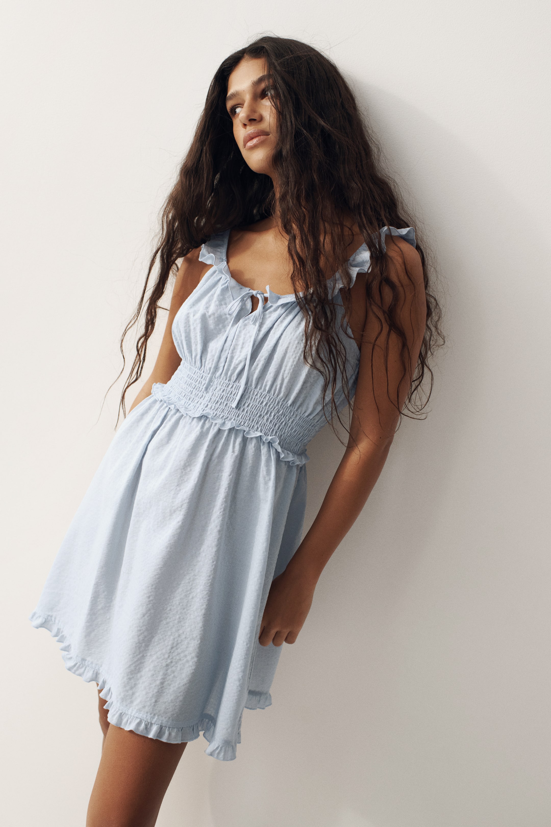 Ampliar la imagen: Se muestra un vestido mini texturizado de color azul claro desde un ángulo de tres cuartos, con tirantes con volantes y un escote cuadrado con detalle de lazo. El vestido presenta un corpiño fruncido y una cintura elástica, que fluye hacia una falda acampanada con un dobladillo con volantes.