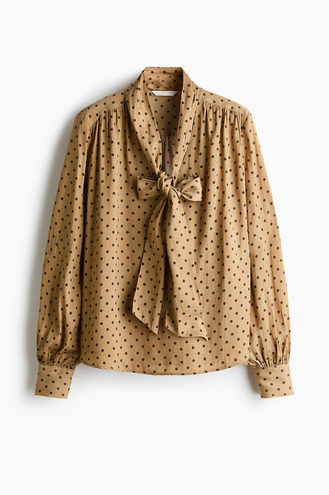 A tan polka-dot blouse is shown facing front, featuring a prominent tie-neck bow at the collar. The blouse is displaying long, slightly voluminous sleeves with gathered shoulders and wide, buttoned cuffs. - 1