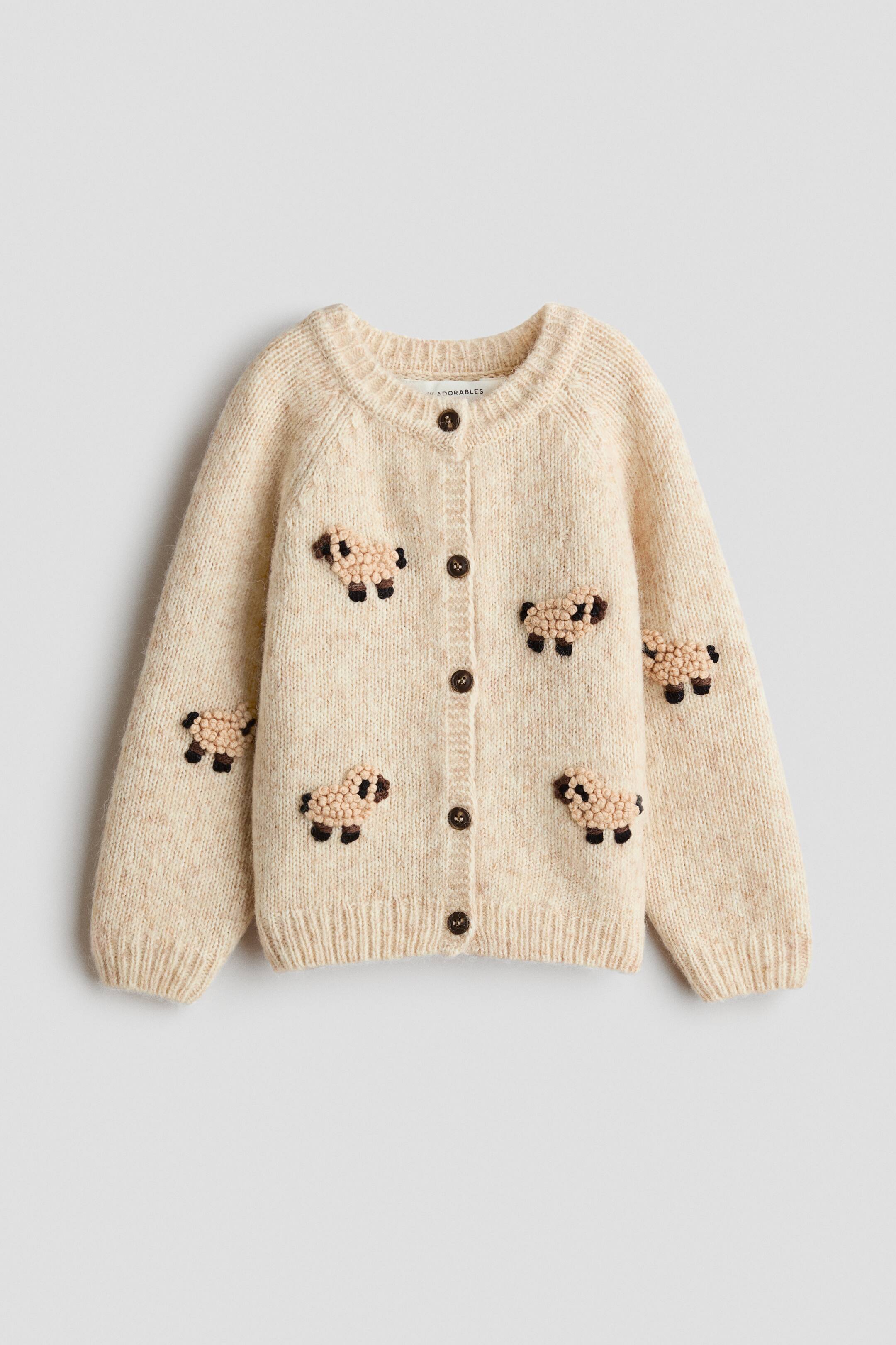 View larger image: A cream-coloured knit cardigan is shown from the front, featuring a ribbed crew neck, cuffs, and hem, along with a button-front fastening with five dark brown buttons. The cardigan displays multiple small, textured sheep motifs with boucle-style bodies and dark brown embroidered details scattered across the front and sleeves.