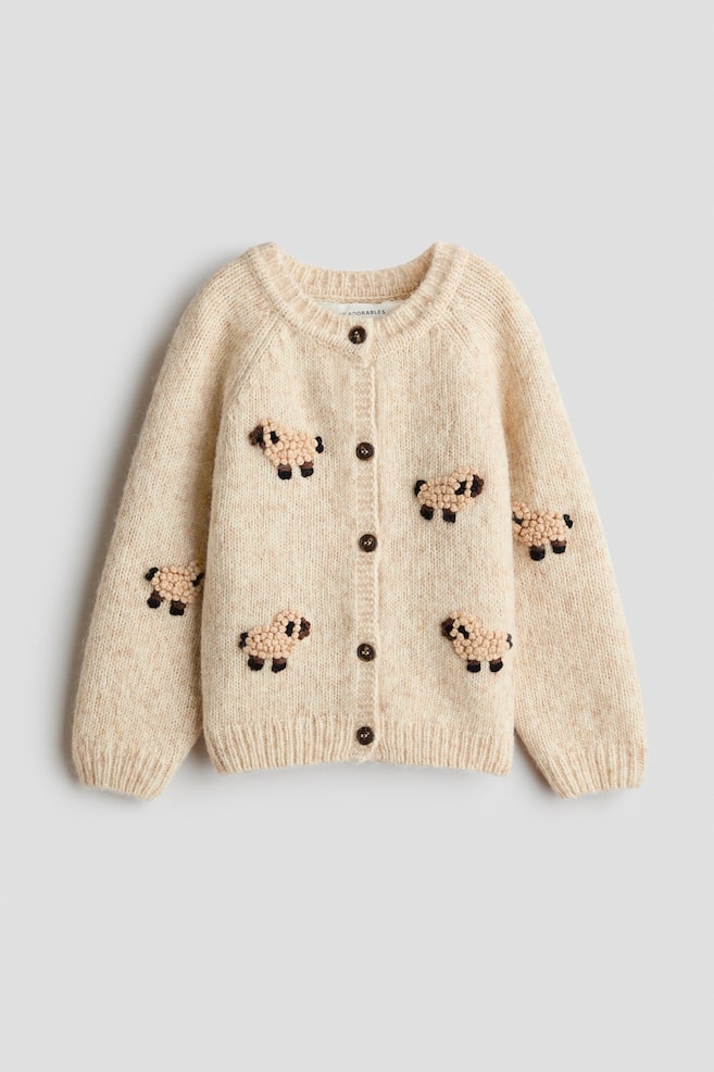 A cream-coloured knit cardigan is shown from the front, featuring a ribbed crew neck, cuffs, and hem, along with a button-front fastening with five dark brown buttons. The cardigan displays multiple small, textured sheep motifs with boucle-style bodies and dark brown embroidered details scattered across the front and sleeves. - 1