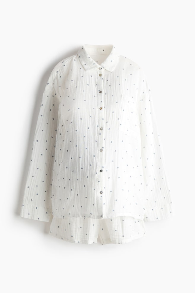 A white pyjama set is presented from the front, consisting of a shirt and shorts in a crinkled fabric patterned with small grey hearts. The shirt features a collar, buttons down the front, a patch pocket on the chest, dropped shoulders and long sleeves; the matching shorts appear below. - 1