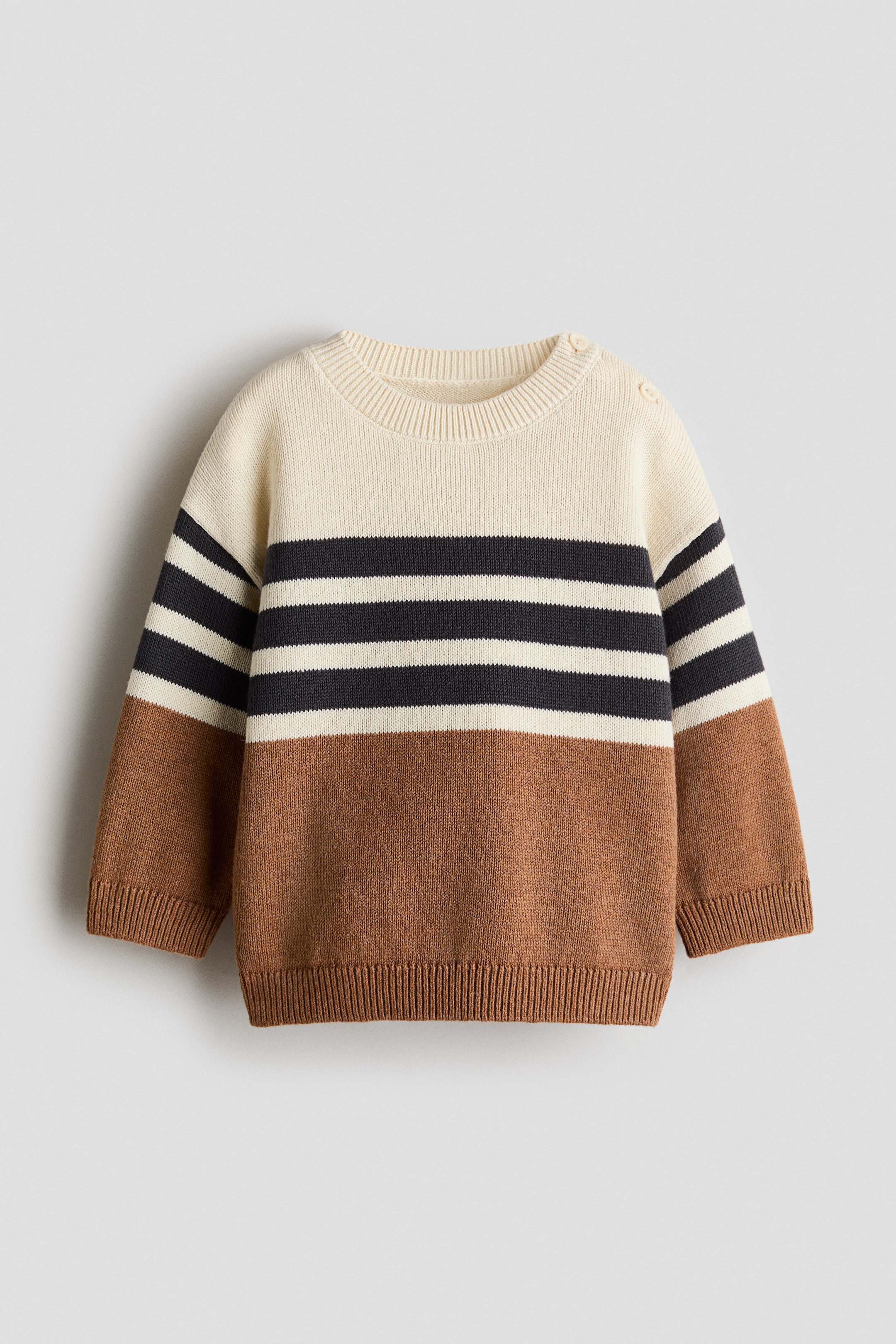 Agrandir l'image: Un pull en maille est posé à plat, présentant une partie supérieure crème avec deux bandes horizontales gris foncé sur la poitrine et les manches, se prolongeant vers une partie inférieure marron unie. Il comporte un col rond côtelé, deux boutons sur l'épaule gauche, ainsi que des poignets et un ourlet côtelés.