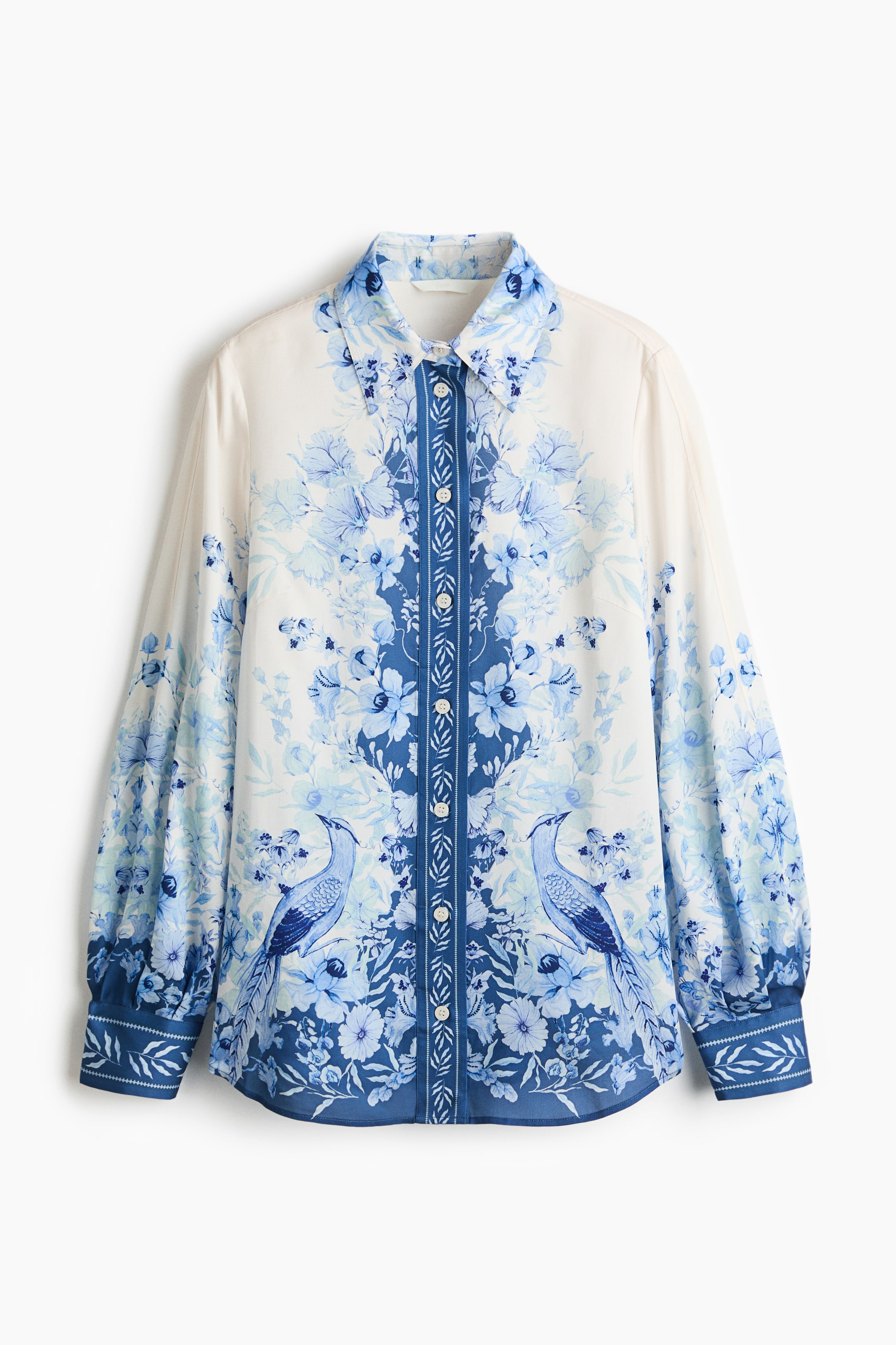 Ampliar la imagen: Una camisa blanca muestra un patrón simétrico de flores y aves azules en su frente. Cuenta con un cuello en punta, una tapeta de botones completa con una franja central con patrón azul, y mangas largas que se recogen en puños anchos con patrón. Dos pájaros azules se enfrentan en la parte inferior del frente.