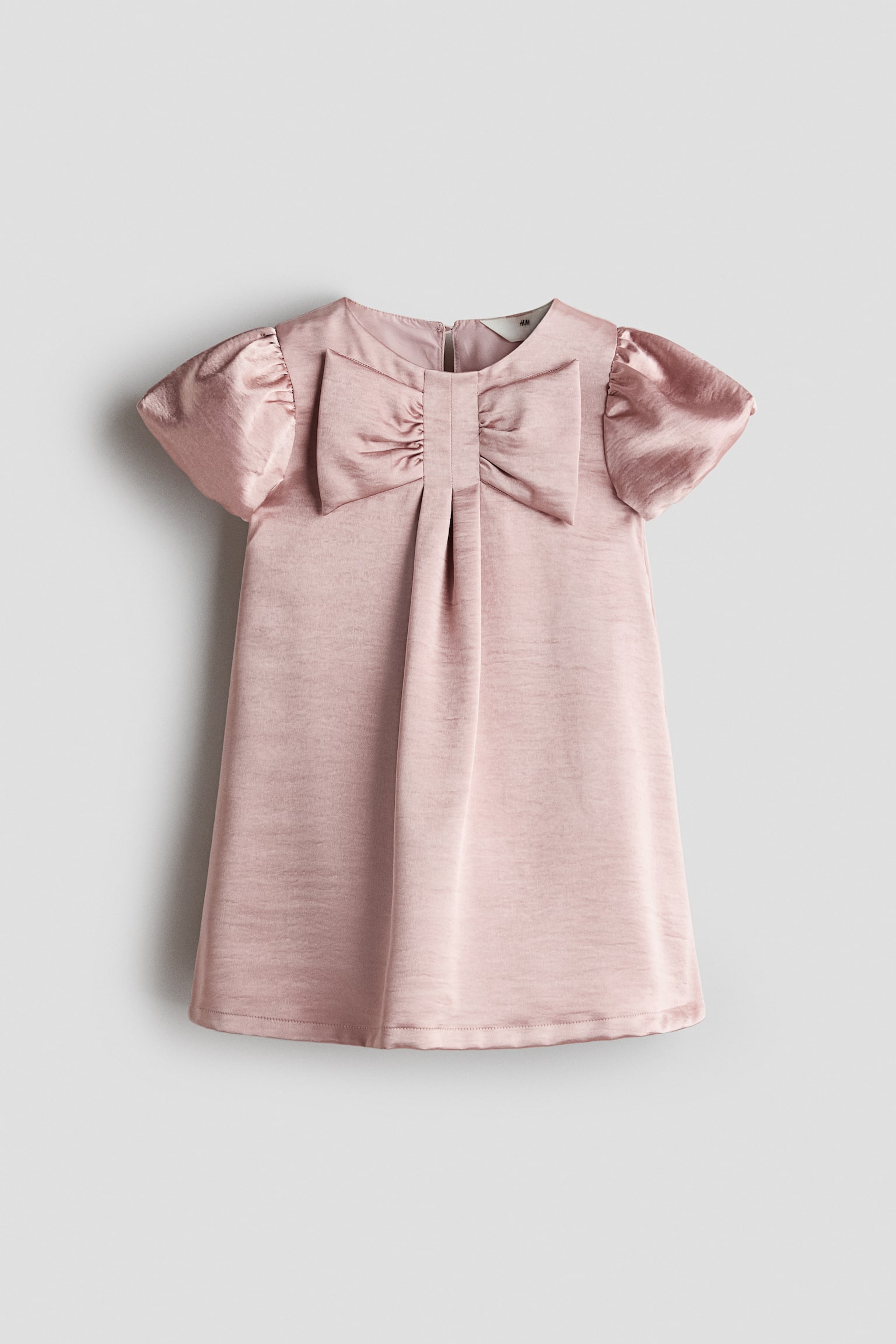 A dusty pink satin-finish dress is shown facing front, featuring a round neckline, short puff sleeves, and a large decorative bow at the centre of the chest. A single pleat runs down the front from beneath the bow to the hem of the A-line skirt.