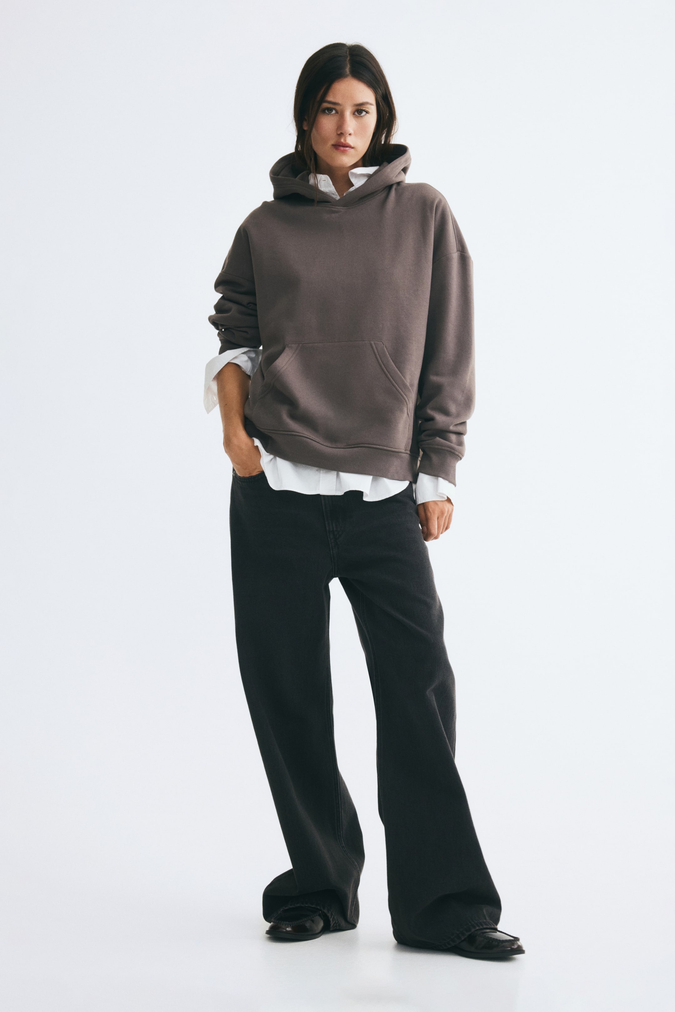 View larger image: The model is wearing a dark gray-brown sweat fabric hoodie layered over a white collared shirt and paired with dark wide-leg jeans. The hoodie features a double-layer hood, low dropped shoulders, a front kangaroo pocket, and ribbing at the cuffs and hem.