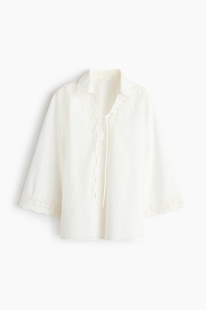 The white top is featuring a spread collar and a V-neckline with a tie detail. It is showcasing broderie anglaise trim around the neckline and the hems of its three-quarter length sleeves, presenting a relaxed fit from the front. - 1
