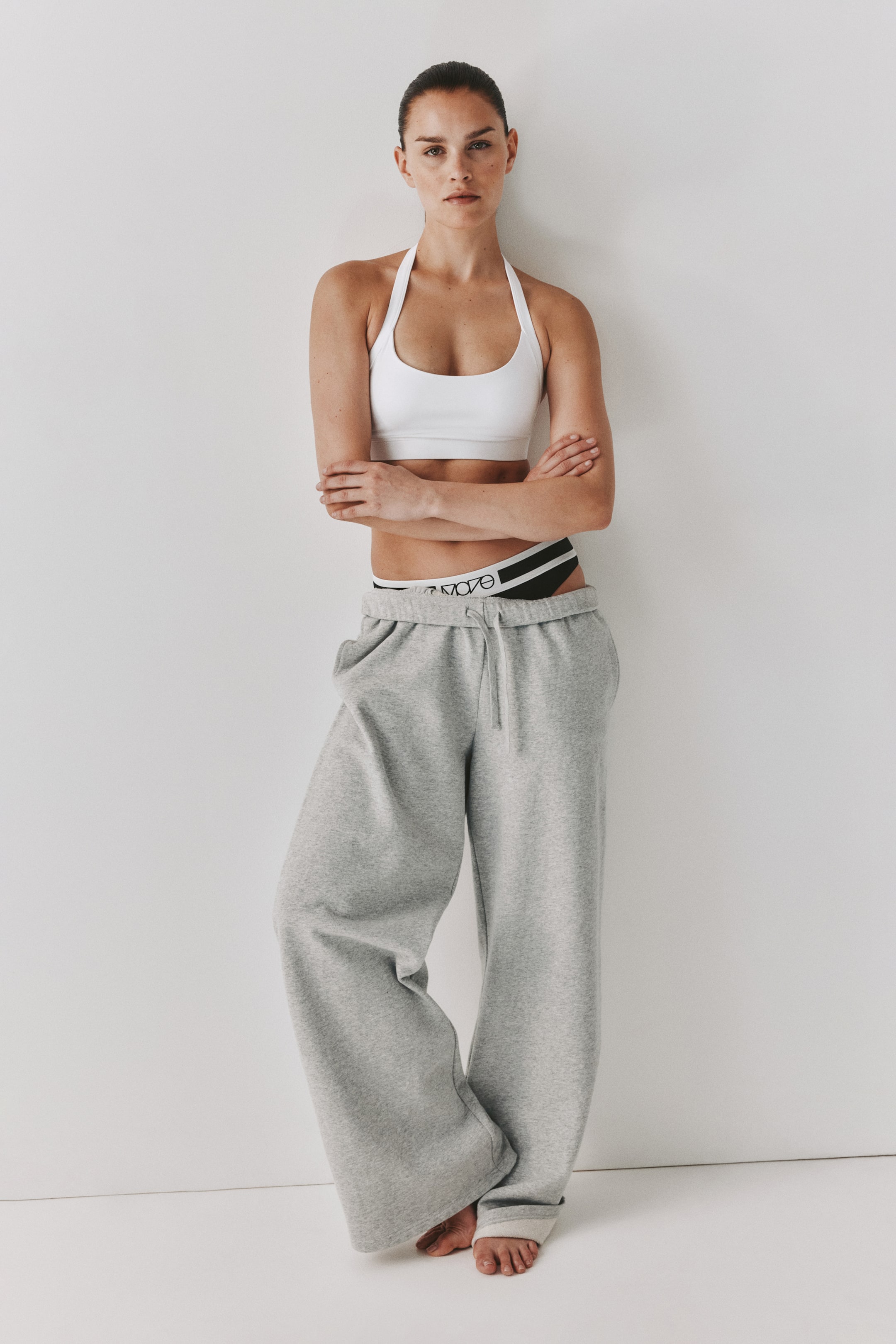 Agrandir l'image: Un soutien-gorge de sport blanc à col licou présente un design à encolure dégagée, tandis qu'un pantalon de survêtement gris à jambes larges met en avant une taille à cordon de serrage et une coupe décontractée. Une ceinture de sous-vêtement rayée noire et blanche avec texte est révélée au-dessus du pantalon de survêtement.
