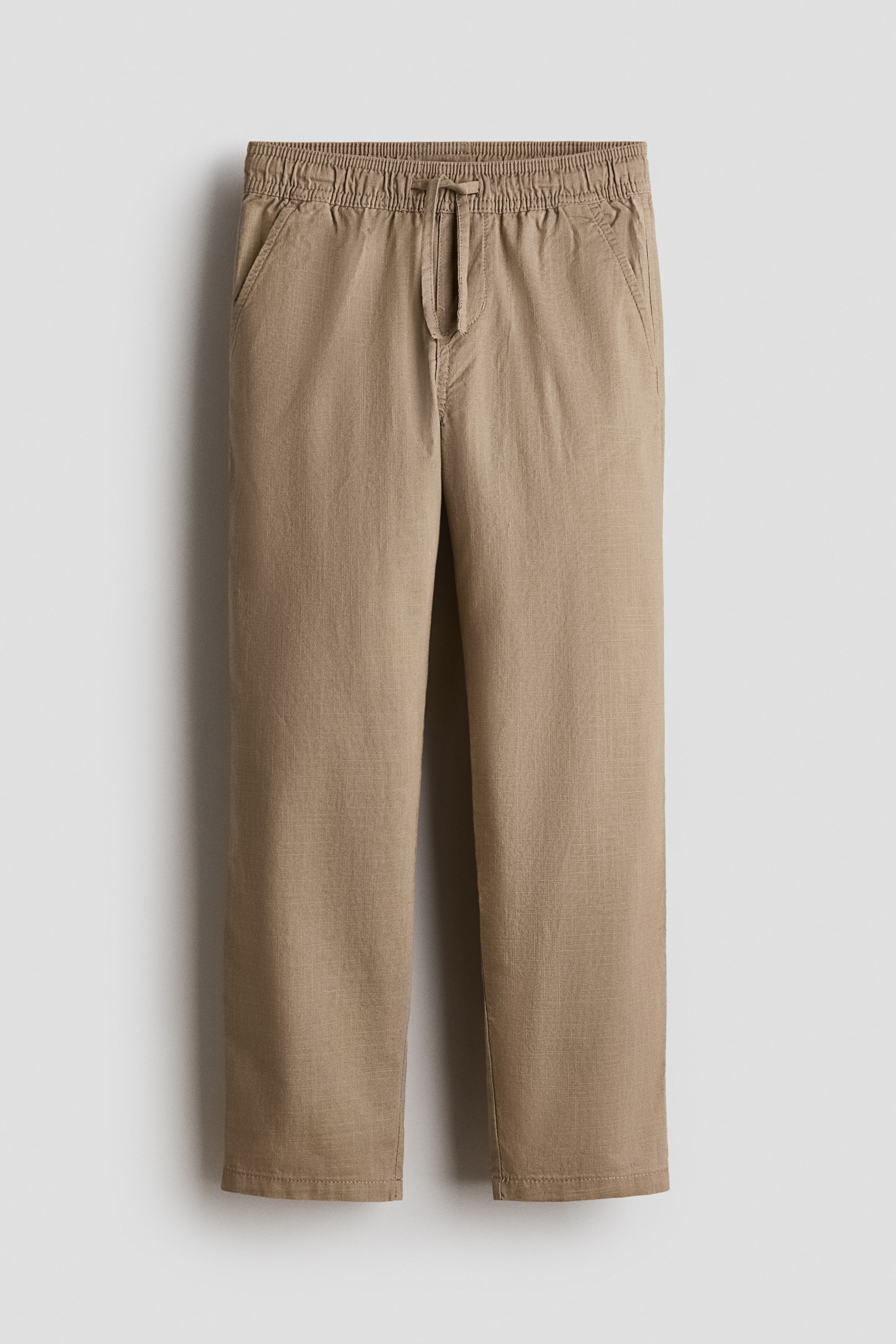 View larger image: The light brown pants feature an elasticized waistband with a visible drawstring, and side slash pockets at the hips. The pants offer a relaxed, straight-leg fit with a subtle woven texture.