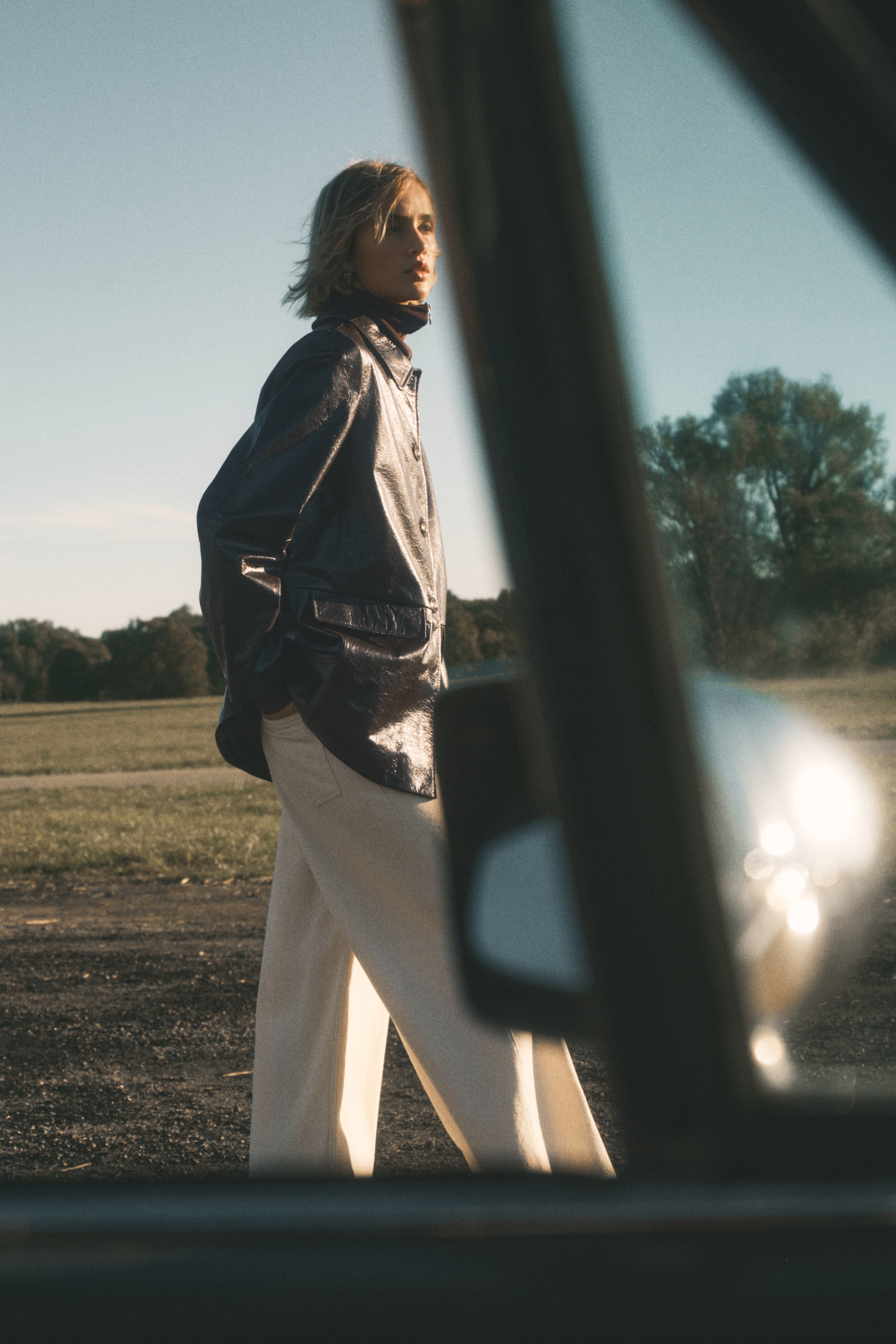 Ingrandisci l'immagine: Una giacca con finitura a effetto vernice increspata in tonalità scura viene indossata, inclinata leggermente lontano dall'osservatore, sopra un top a collo alto scuro. La giacca è caratterizzata da un colletto a punta, una chiusura frontale con bottoni di colore chiaro e grandi tasche con pattina, ed è abbinata a pantaloni ampi di colore chiaro.