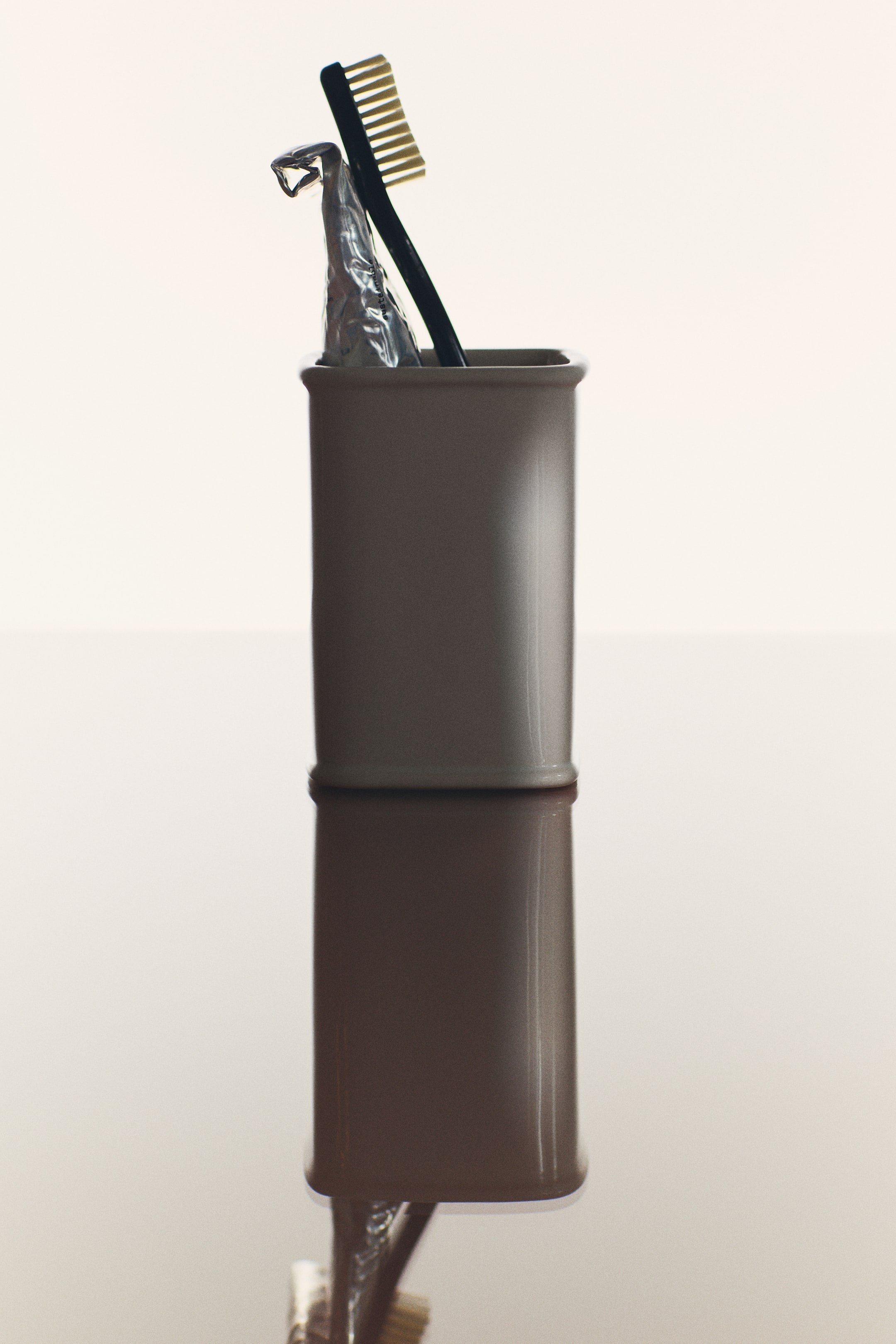 View larger image: A beige stoneware toothbrush holder with a glossy finish and a rectangular design with rounded contours is standing front-facing, holding a black toothbrush and a silver toothpaste tube. The container and its contents are reflected onto a glossy surface below.