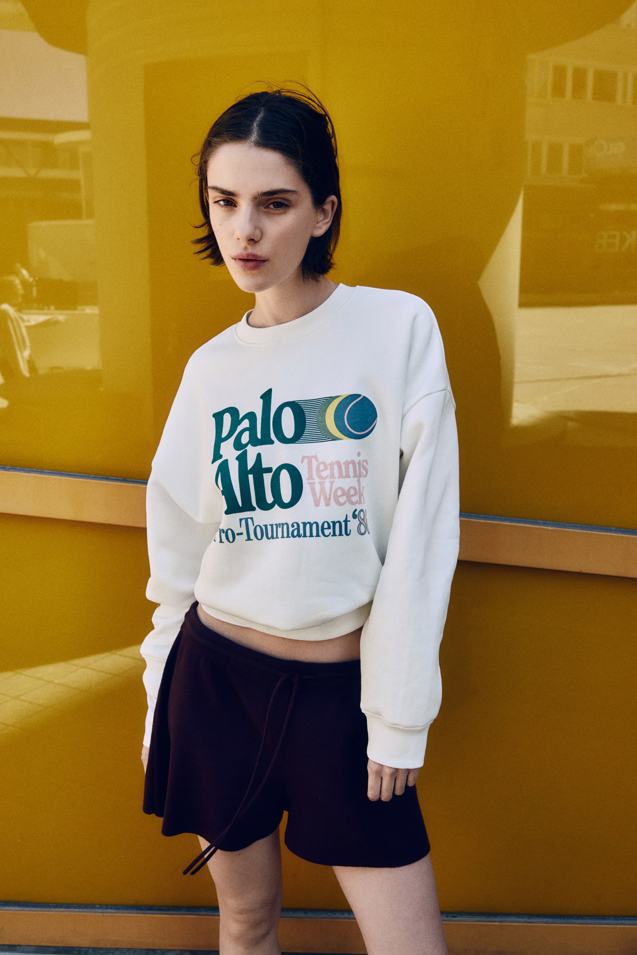 View larger image: An off-white oversized top in printed sweatshirt fabric, featuring a "Palo Alto Tennis Week Pro-Tournament '88" graphic, is being worn by the model. It has a round, rib-trimmed neckline, dropped shoulders, long sleeves with ribbed cuffs, and a ribbed hem that is positioned above the waist. Dark shorts are being worn below the top.
