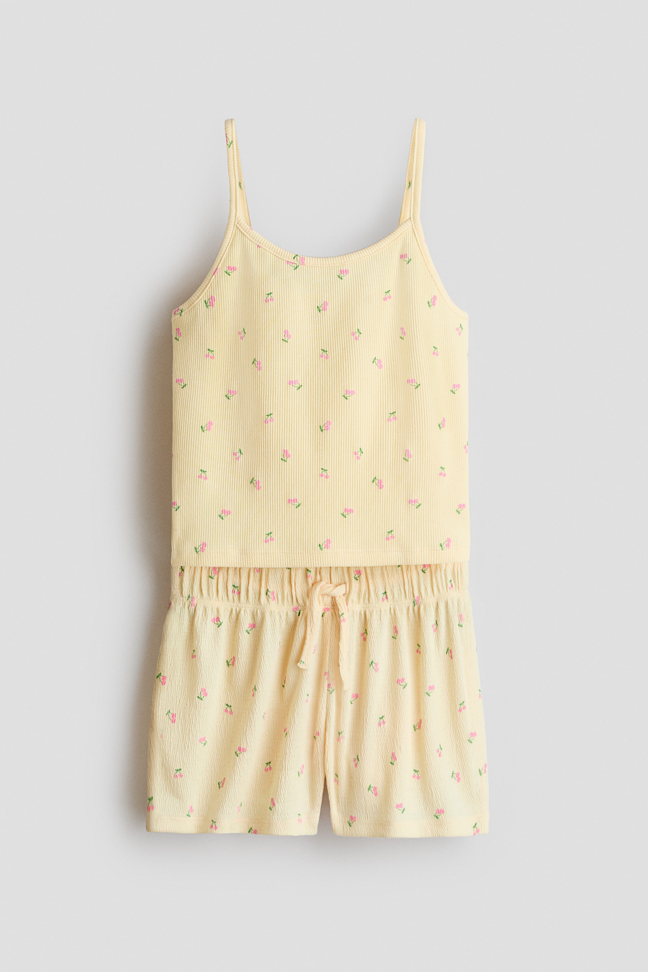 Ver imagem maior: Um conjunto de pijama de duas peças em amarelo claro é apresentado de frente, com um top de alças finas canelado e calções texturizados a condizer. Ambas as peças são decoradas com motivos de cerejas cor-de-rosa dispersas, e os calções incluem uma cintura elástica com cordão ajustável.