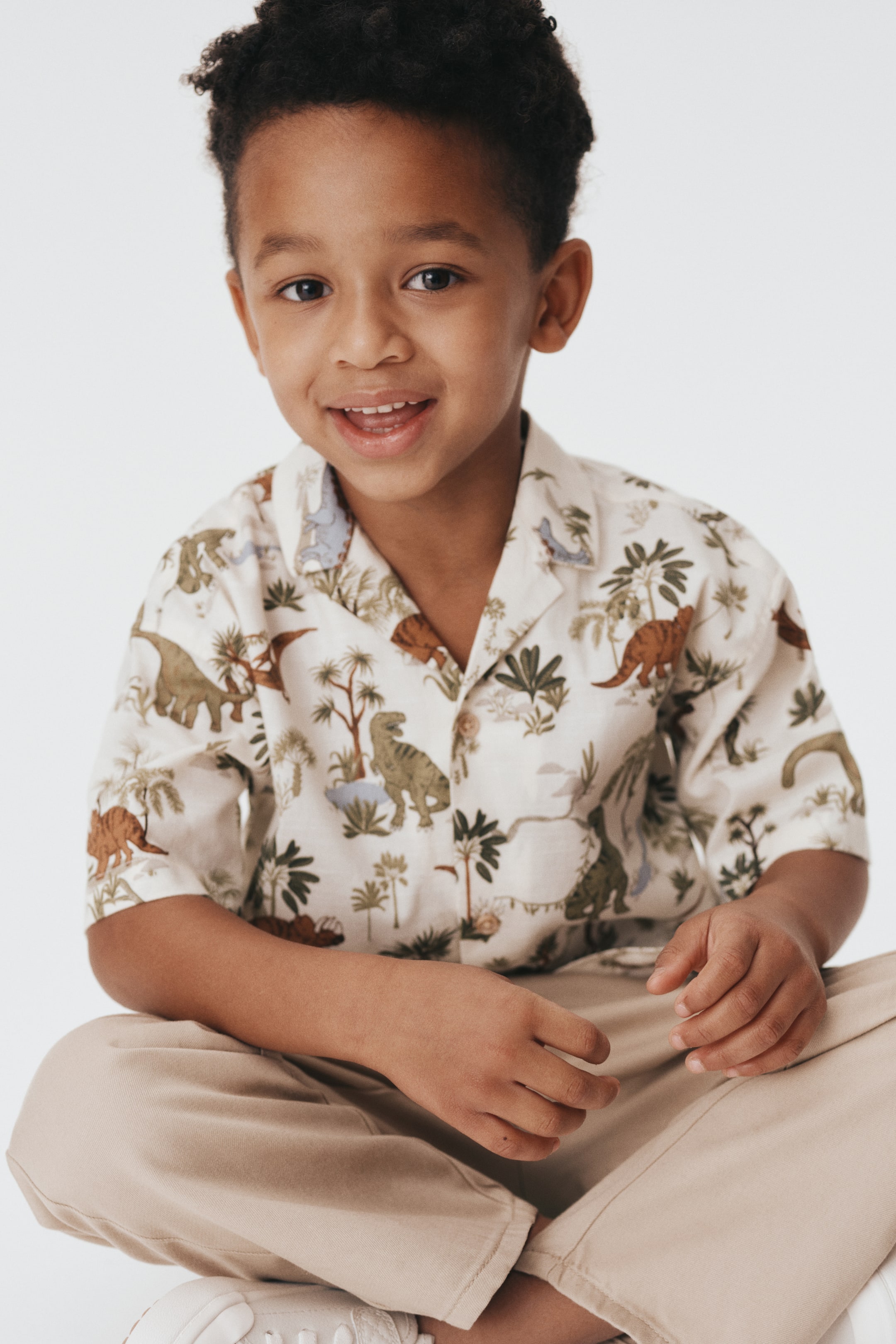 View larger image: A short-sleeved, off-white button-up shirt is being presented front-facing, featuring a brown and green dinosaur and palm tree print. The shirt has an open Cuban collar and light-colored buttons, and is being styled with beige pants.