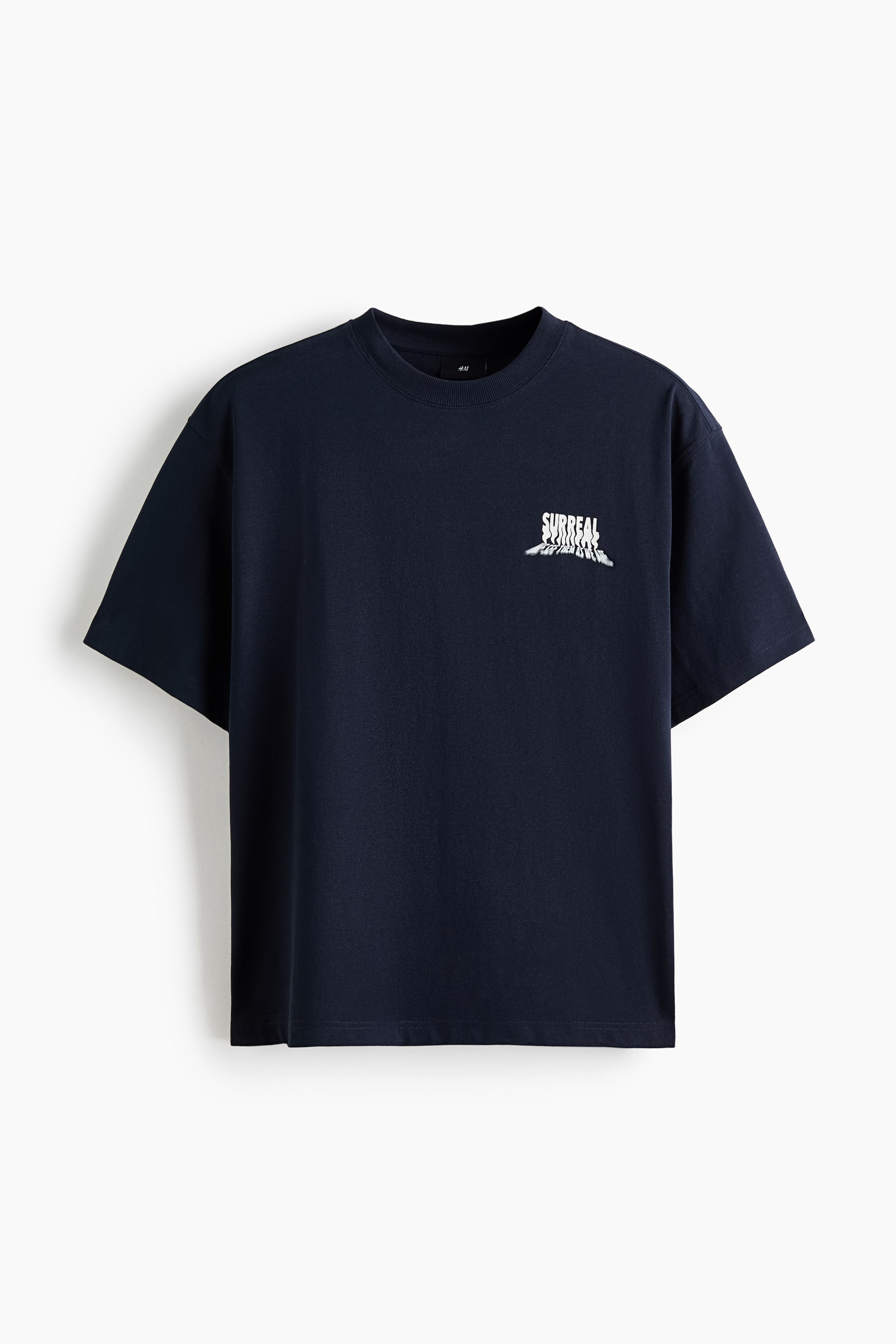 拡大画像を表示: 濃紺のTシャツが平らに置かれており、リブ仕様のクルーネックと半袖が見えます。左胸には、歪んだ波打つような効果のある「SURREAL」という文字と、その下に小さなテキストを配した白いグラフィックプリントが施されています。