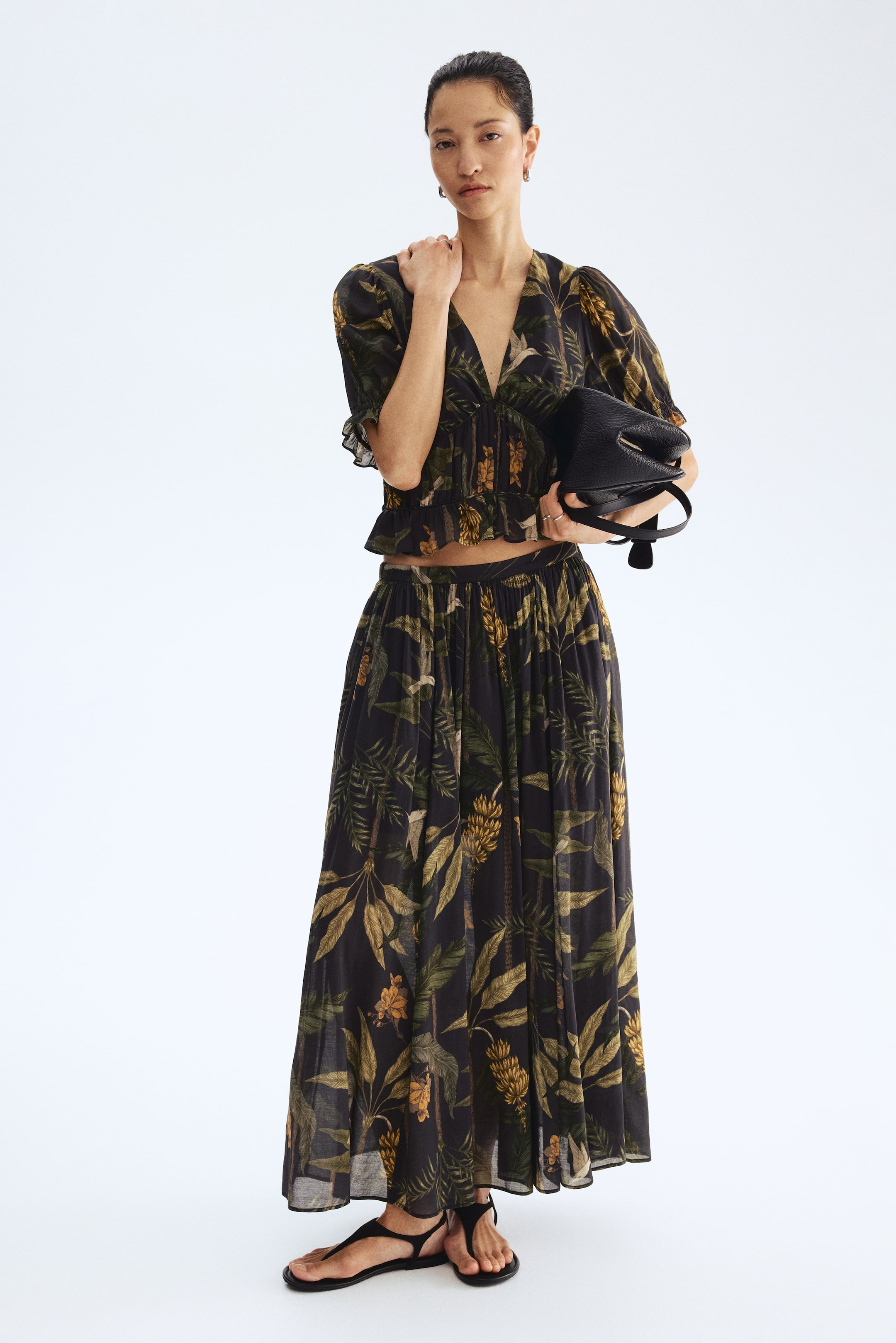 הצגת תמונה גדולה יותר: סט חולצת קרופ תואמת וחצאית מקסי מציג רקע כהה עם דפוסי עלים ופירות טרופיים בירוק וצהוב, מוצג כלפי קדימה. החולצה בעלת מחשוף V מציגה שרוולים נפוחים קצרים ושוליים מקופלים, בעוד החצאית הזורמת נמשכת עד הקרסוליים. תיק יד שחור עם טקסטורה מוחזק, וסנדלים שחורים עם רצועות משלימים את המראה.