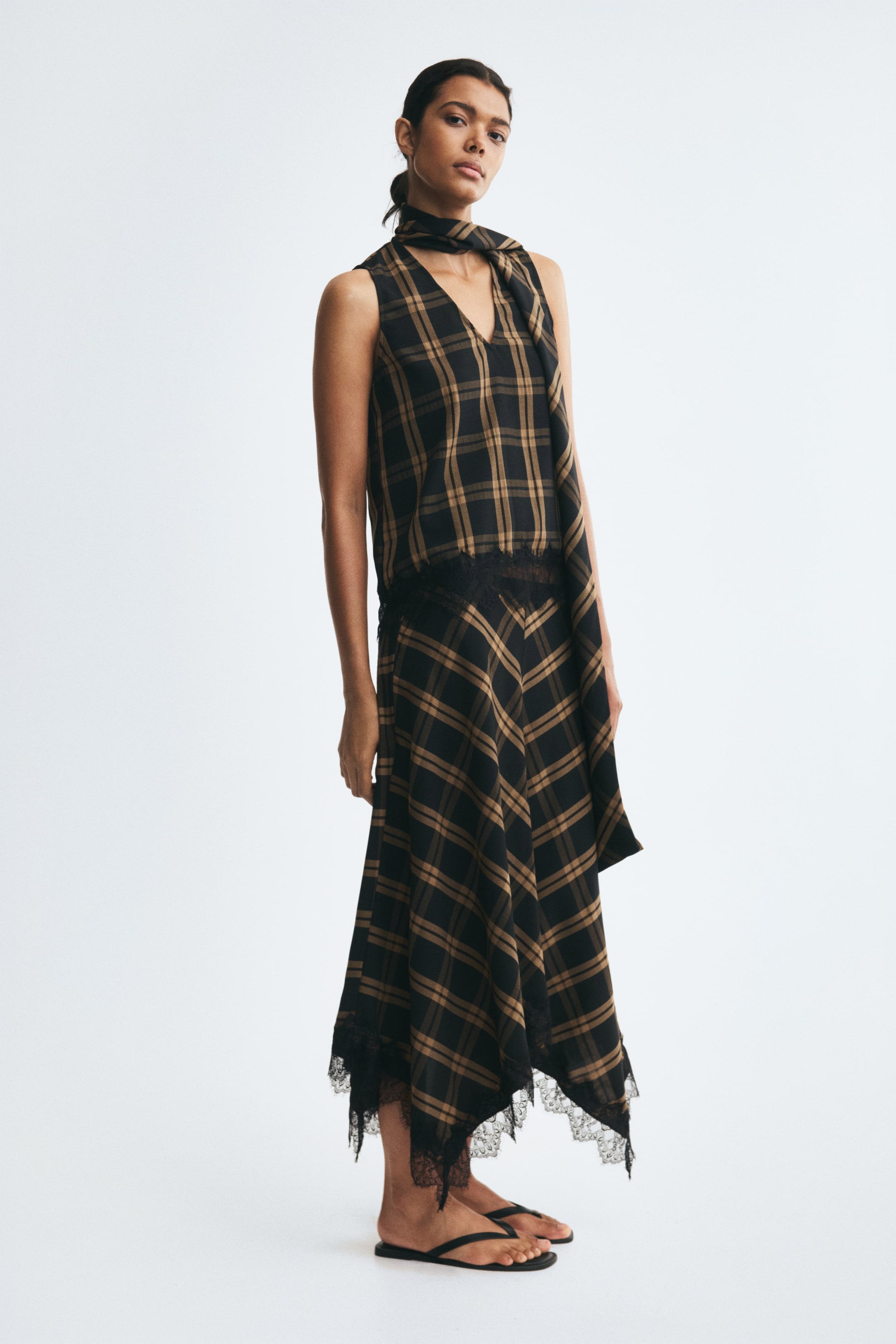 View larger image: A loose-fit maxi skirt in a brown and black plaid pattern is being worn by the model, featuring a handkerchief hem finished with black lace trim. The skirt is styled with a matching sleeveless plaid top and flat black sandals.