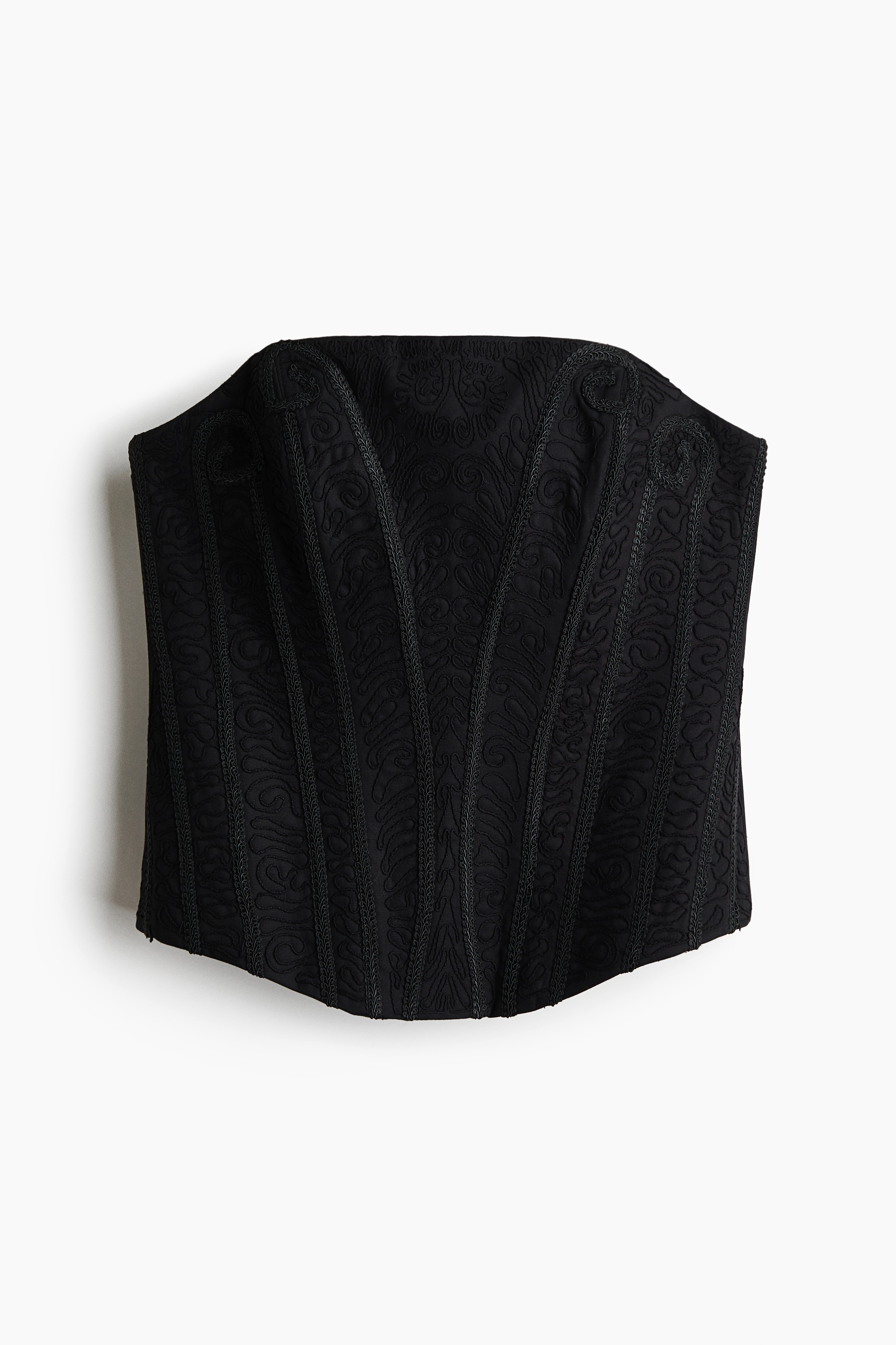 Un haut corset bandeau noir, ornÃ© dâune broderie ton sur ton raffinÃ©e et de baleines verticales structurÃ©es, est portÃ© par le mannequin. Le haut bustier est superposÃ© Ã  une blouse blanche Ã  col montant, dotÃ©e de manches bouffantes volumineuses, et associÃ© Ã  un pantalon noir.