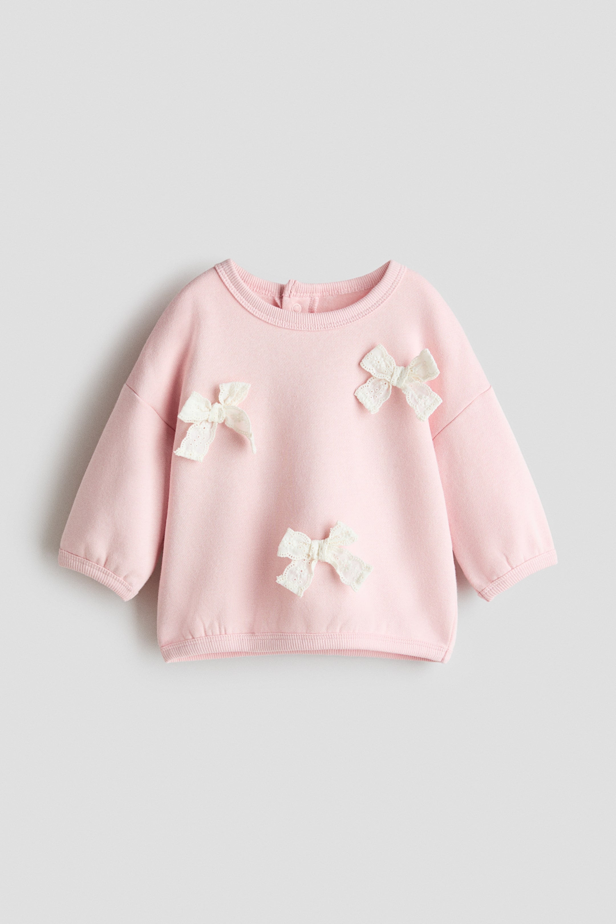 View larger image: A pink sweatshirt top is facing FRONT, featuring a round, rib-trimmed neckline with press-studs visible at the back. It is adorned with three cream-coloured lace bows on the front, and is designed with low, dropped shoulders and ribbed cuffs and hem.