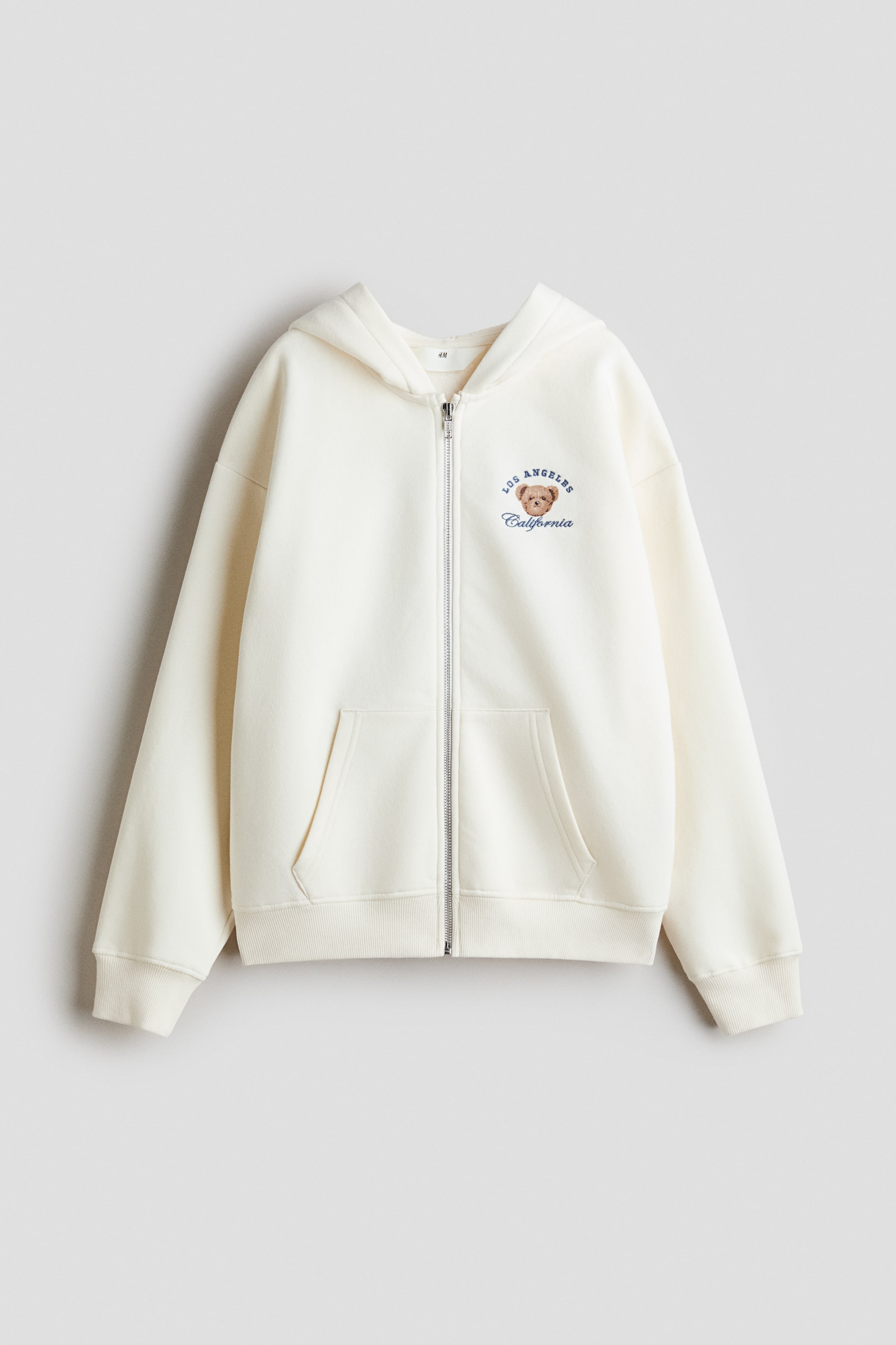 Ampliar la imagen: Una sudadera con capucha color blanco roto, de corte oversize y cierre frontal, se muestra de frente: cuenta con una cremallera central y un motivo bordado en el pecho izquierdo que representa la cabeza de un oso marrón con el texto 'LOS ANGELES California'. Está diseñada con hombros caídos, capucha forrada en jersey, bolsillos tipo canguro frontales y puños y dobladillo acanalados.