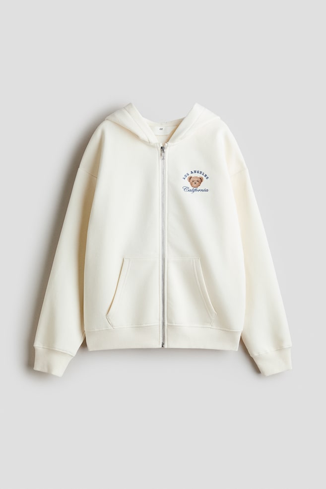 An off-white oversized zip-through hoodie is presenting its FRONT, featuring a central zipper and an embroidered motif on the left chest depicting a brown bear head with 'LOS ANGELES California' text. It is designed with dropped shoulders, a jersey-lined hood, front kangaroo pockets, and ribbed cuffs and hem. - 1