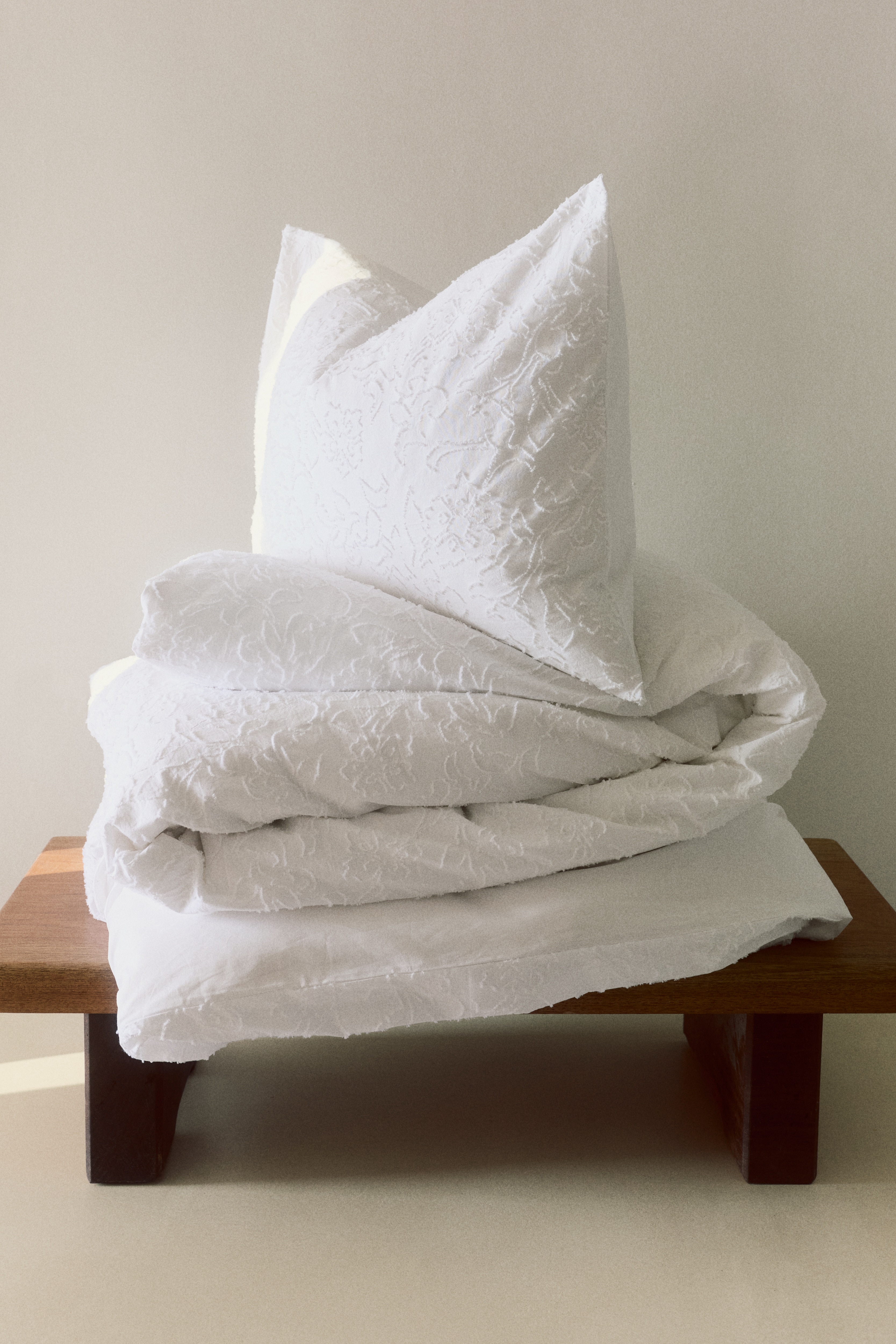 Un juego de cama blanco presenta una funda de almohada y dos fundas de edredón dobladas, apiladas verticalmente. Cada artículo muestra un sutil patrón floral texturizado en toda su superficie.