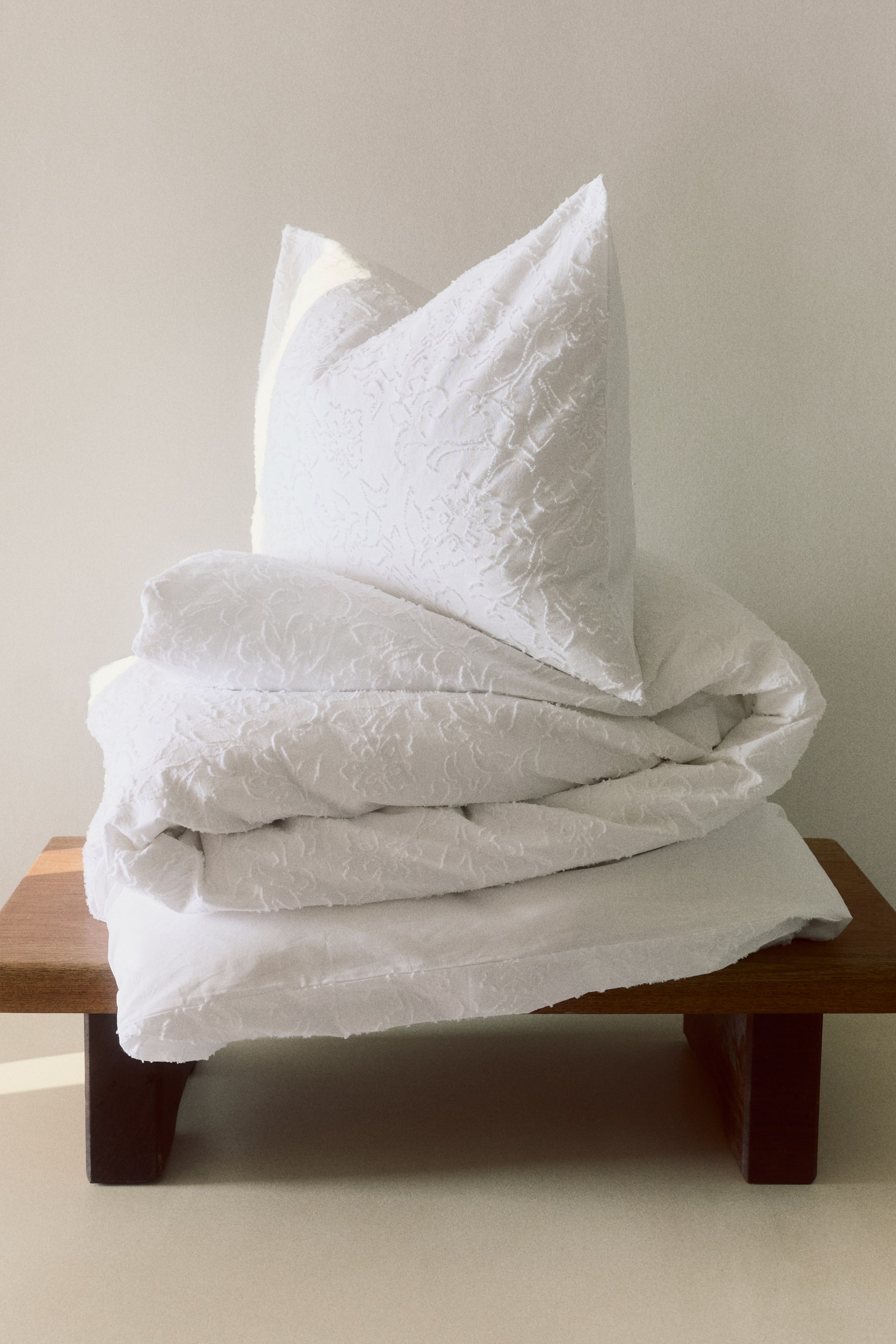 Ampliar la imagen: Un juego de cama blanco está apilado, con una almohada colocada en posición vertical e inclinada hacia la derecha, un edredón doblado y una sábana plana, todos mostrando un patrón texturizado en relieve similar al damasco.