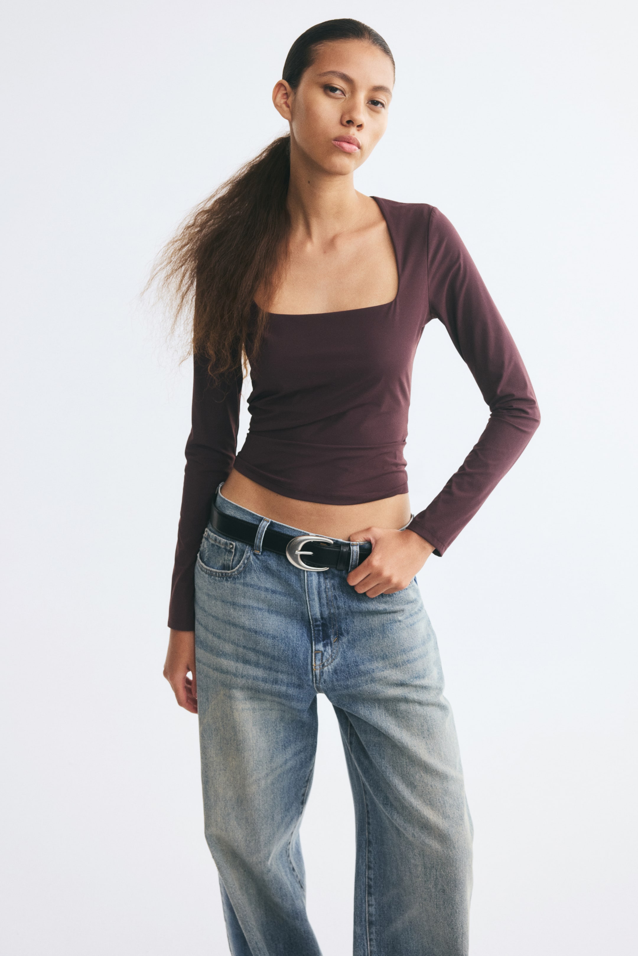 View larger image: A fitted, dark plum-coloured top featuring a deep square neckline and long sleeves is being worn by the model. The top is styled with low-rise blue jeans and a black belt.