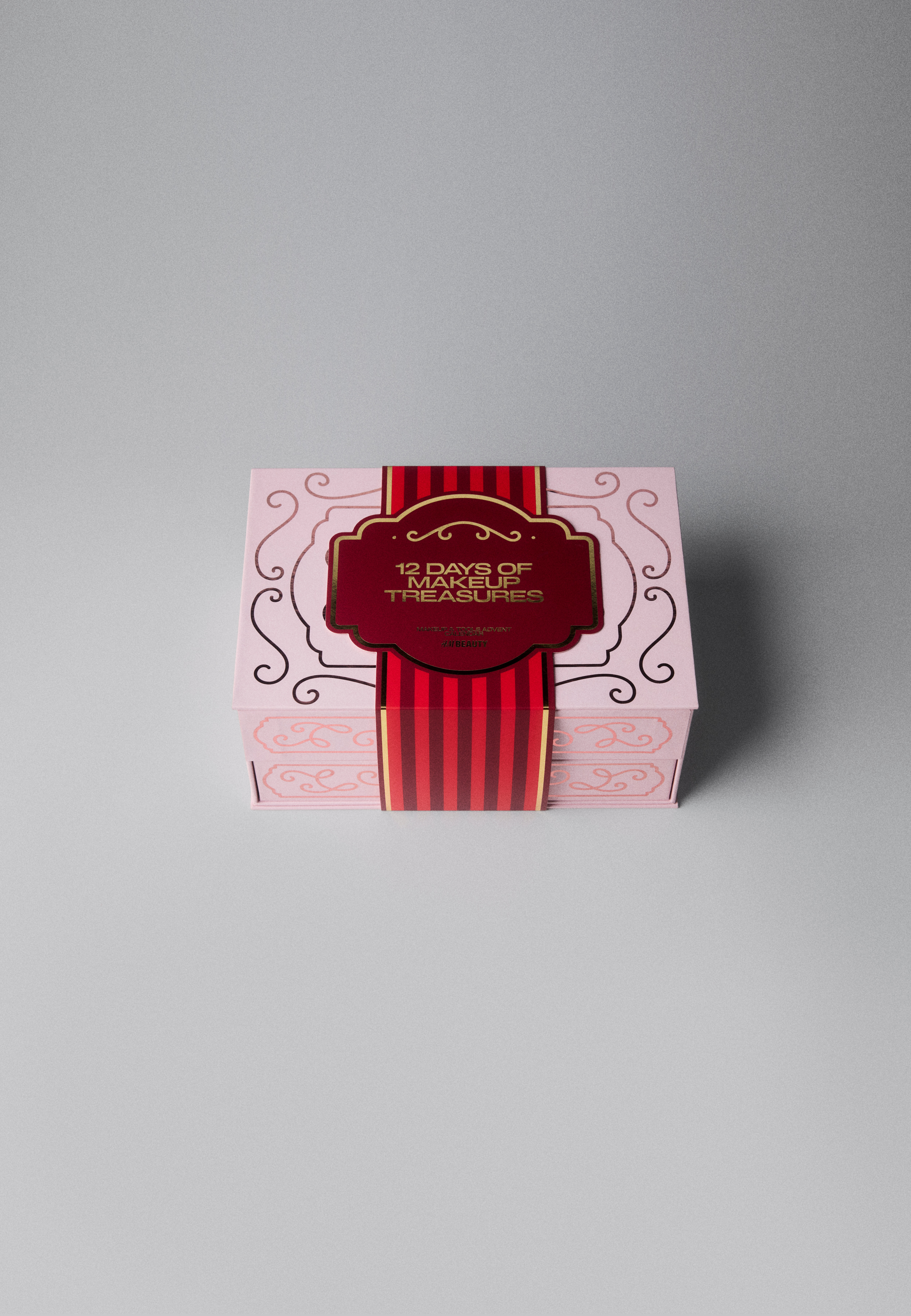 Le calendrier de lâAvent Beauté 12 jours présente un coffret décoratif rose clair, orienté de face, orné de volutes métalliques roses et dâun tiroir inférieur. Quatre petites boîtes-cadeaux numérotées, aux divers motifs et illustrations roses, sont empilées sur le dessus du coffret principal.