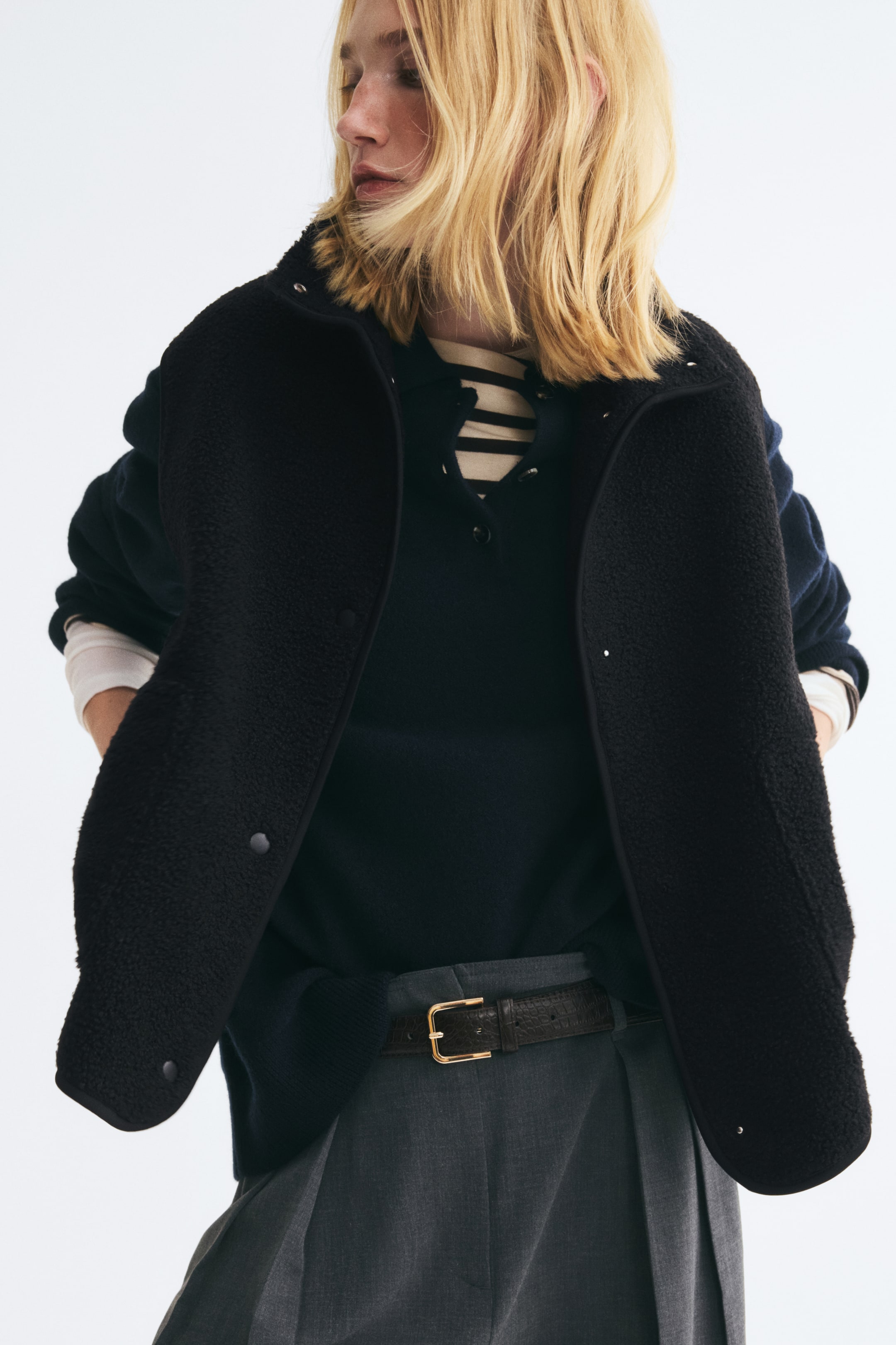 View larger image: A black teddy vest with a funnel collar, jersey binding, and snap-button closures is worn open by the model. The vest is layered over a dark knit polo sweater, a striped top, and gray trousers.