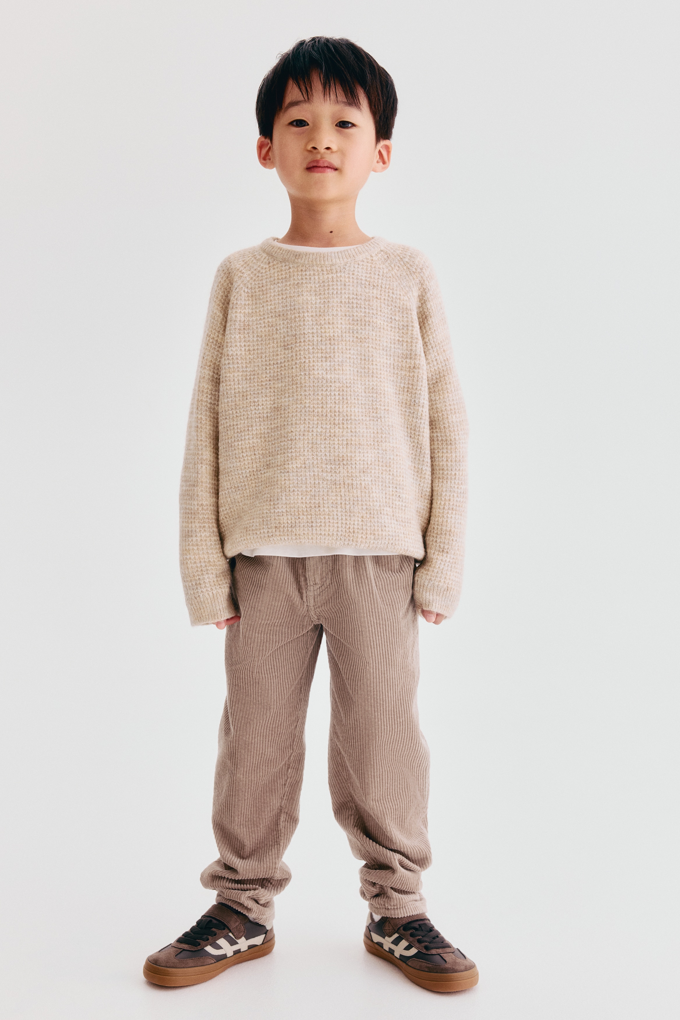 The light beige corduroy trousers are shown from the front, highlighting an elasticated waist with visible gathering and a metal press-stud fastening above a fly seam. They feature two rounded front hip pockets and a small coin pocket, exhibiting a loose fit through the hip, thigh, and leg.