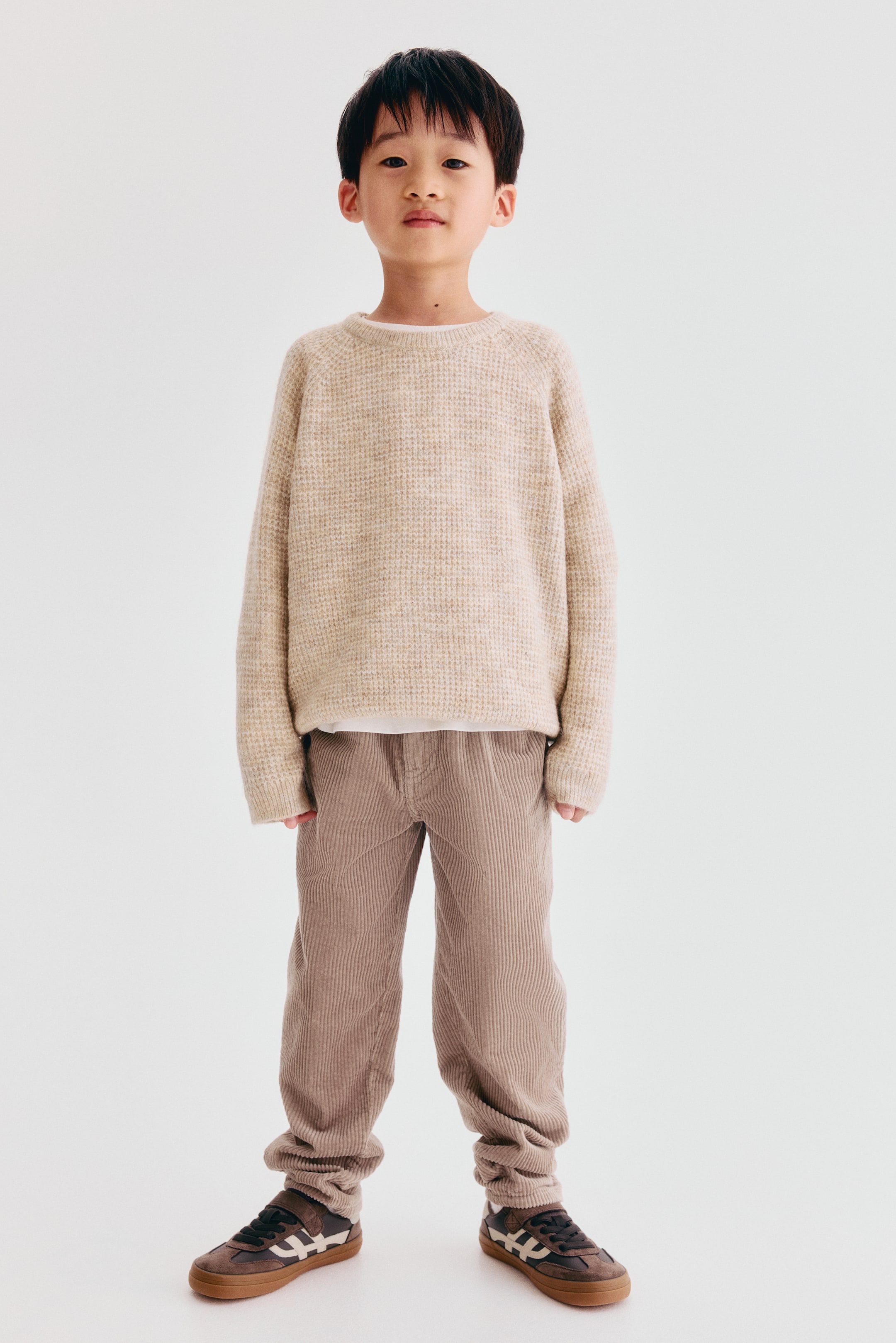 Agrandir l'image: Ce pantalon en velours côtelé de coton brun clair présente une coupe ample au niveau des hanches, des cuisses et des mollets, vu de face. Le pantalon met en valeur un tissu côtelé texturé et se resserre légèrement aux chevilles, au-dessus des baskets.