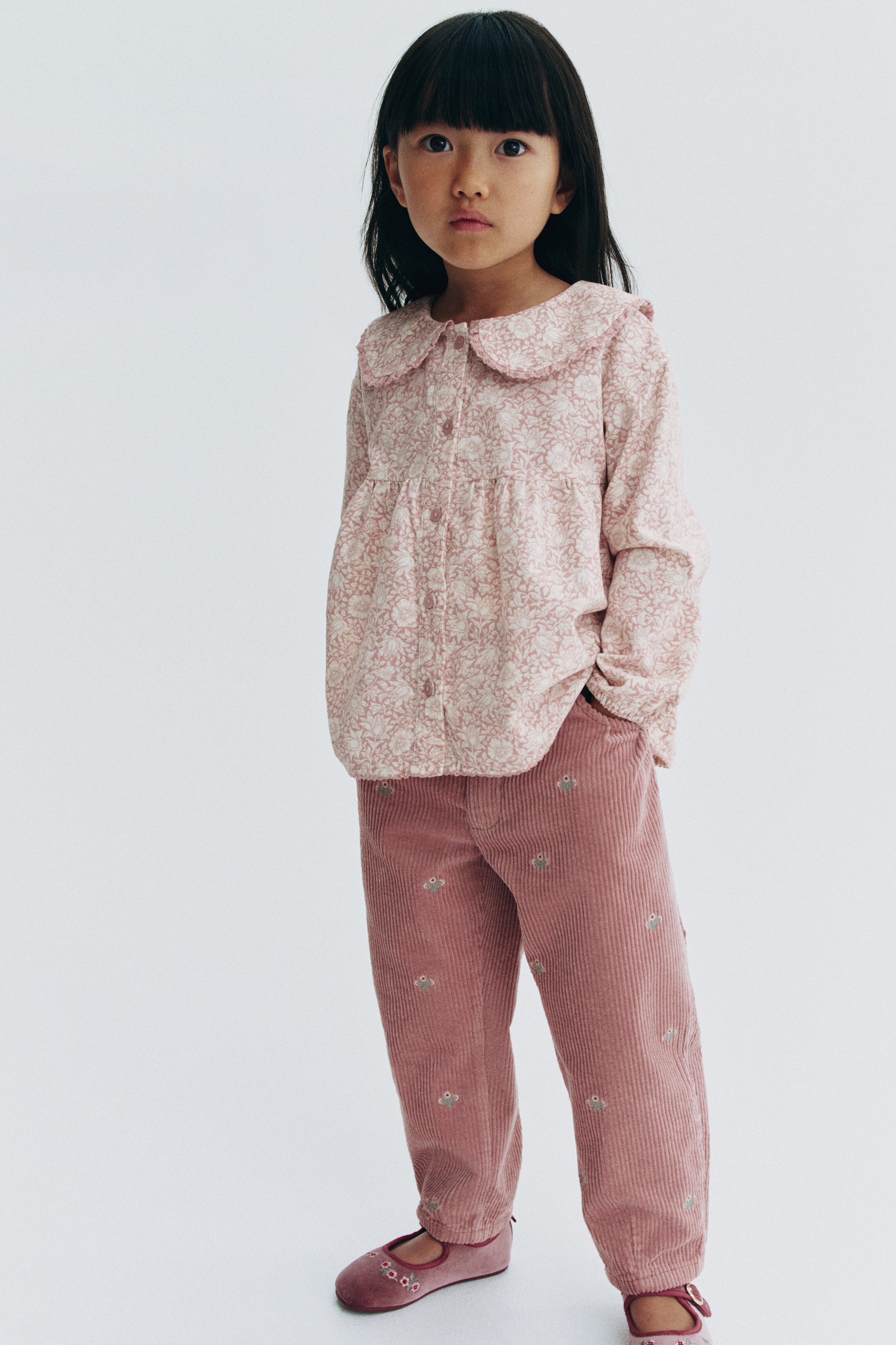 A pair of pink cotton corduroy pants is shown from the front, featuring barrel-style legs with subtle top pleats and small, all-over embroidered floral details. The pants display diagonal front pockets and an elasticated waistband with belt loops and a press-stud closure.
