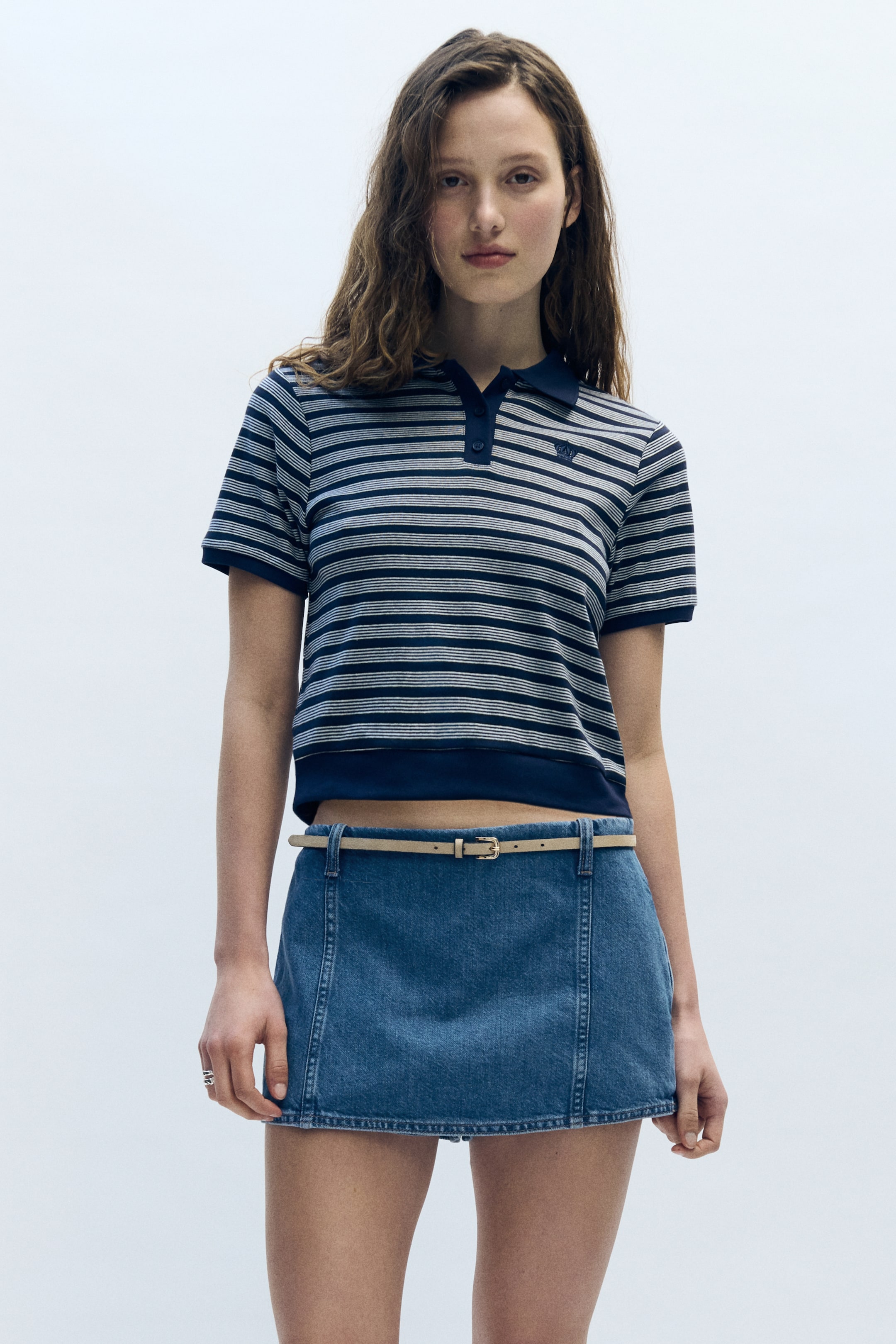 View larger image: "A cropped polo shirt featuring navy blue and white horizontal stripes, a navy blue polo collar, and a two-button placket. A small embroidered logo is visible on the left chest, and the shirt finishes with a solid navy blue band at the hem."