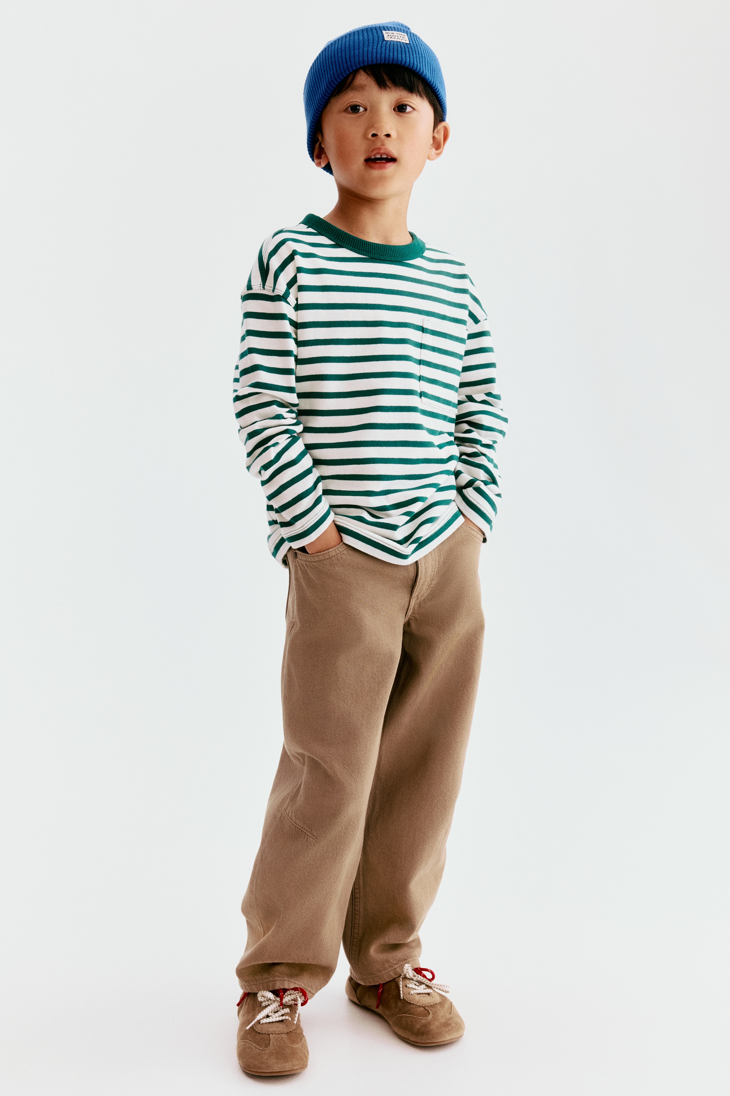 Tan trousers are shown from the front, featuring an elasticated waistband with belt loops and a single button fastening. They also display two front hip pockets and distinctive rectangular knee patches on each wide leg.