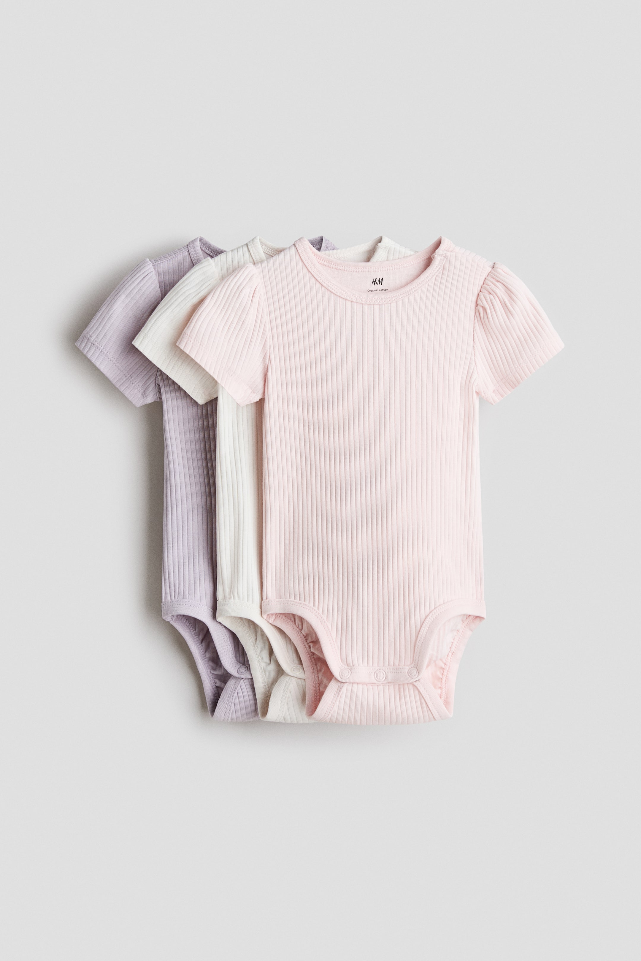 Nagyobb kép megtekintése: A rövid ujjú bodyk puha, bordázott pamutjerseyből készülnek; kerek nyakkivágással, alul patentokkal és a lábak körüli ráncolt varrásokkal. Három body elölről látható: a rózsaszín enyhén puffadt ujjakkal rendelkezik, és a krémszínű illetve lila bodyk fölé van rétegezve.