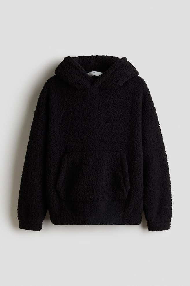 Ein schwarzer Teddy-Hoodie wird von vorne präsentiert, mit einer Kapuze mit überlappender Vorderseite, überschnittenen Schultern, einer Kängurutasche und elastischen Bündchen und Saum. - 1