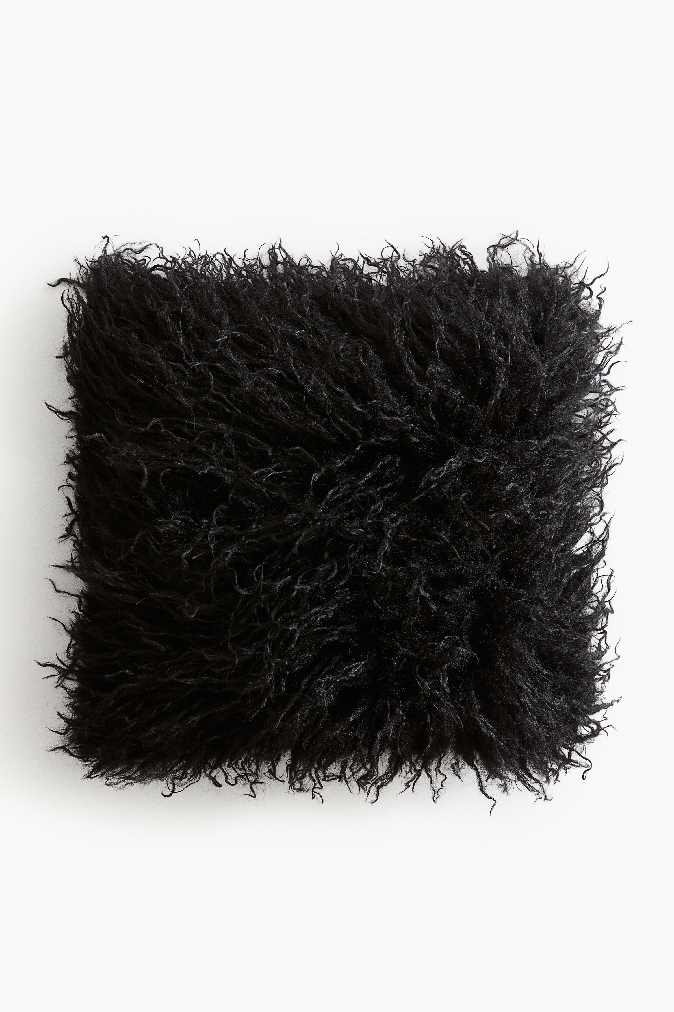 A black square cushion cover featuring long, shaggy, fluffy textured fibres is resting upright on a wooden stool, facing forward. The dense, wavy texture covers the entire surface of the cushion, highlighting its soft and voluminous appearance.