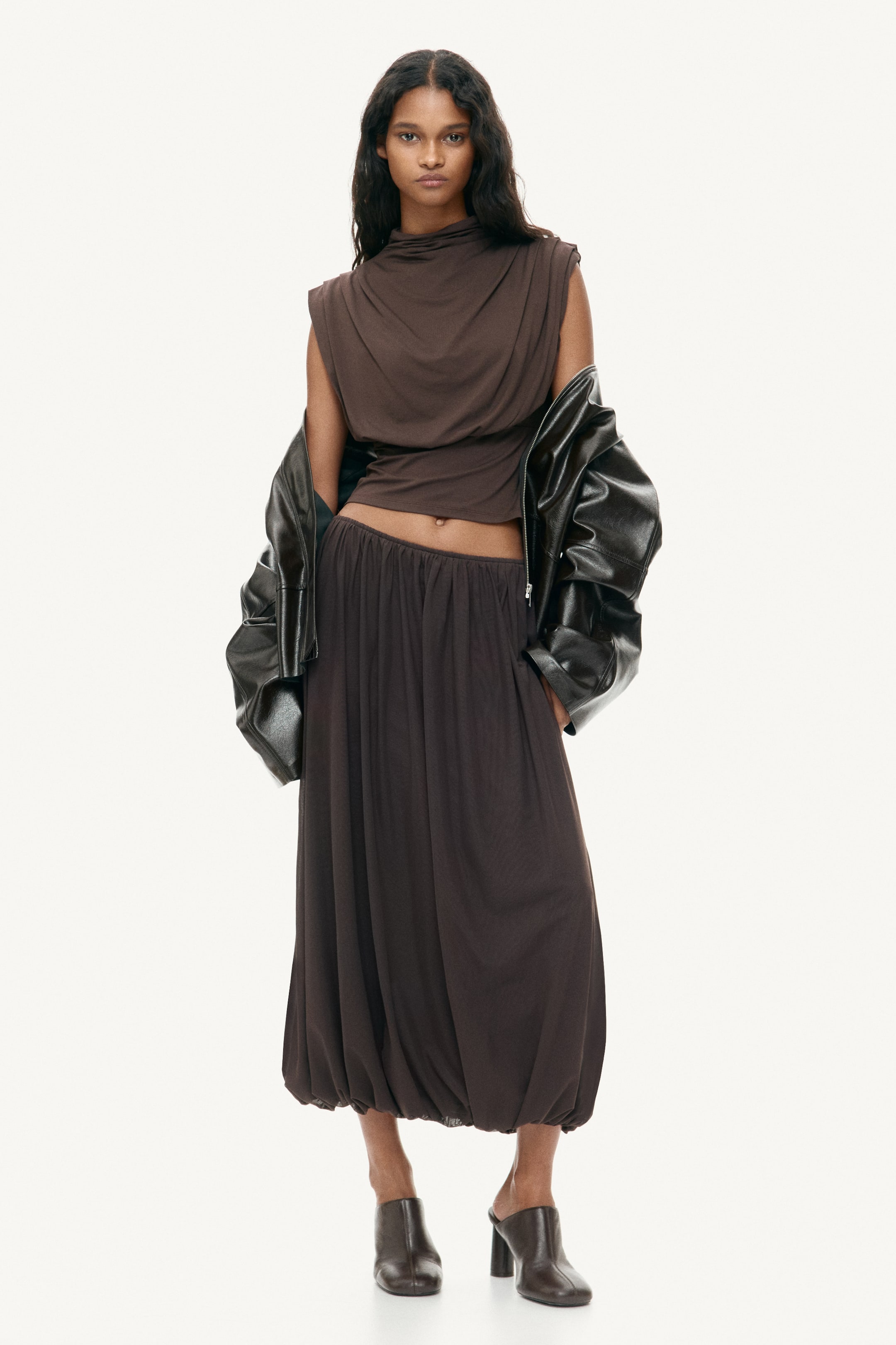 Visualizza immagine più grande: Un top senza maniche marrone scuro con scollo drappeggiato è abbinato a una lunga gonna a palloncino dello stesso colore. Una giacca marrone scuro è appoggiata sulle spalle e dei sabot marrone scuro completano il look.