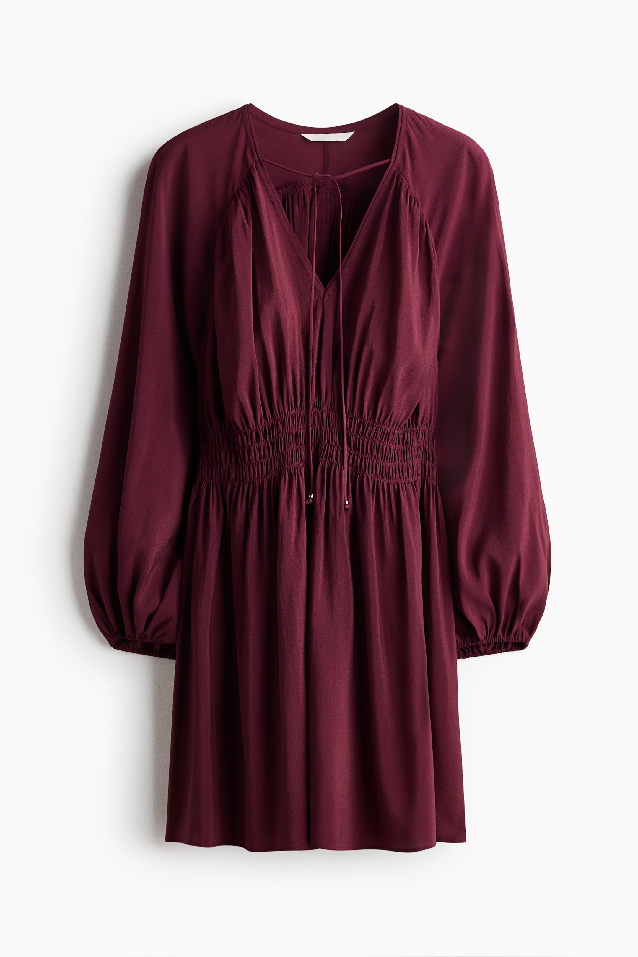 View larger image: A short, oversized maroon dress is displayed from the front, highlighting a deep V-neckline with spaghetti ties and a gathered front yoke. It features long, voluminous balloon sleeves with elasticated cuffs, a smocked section at the waist, and a gently flared skirt.
