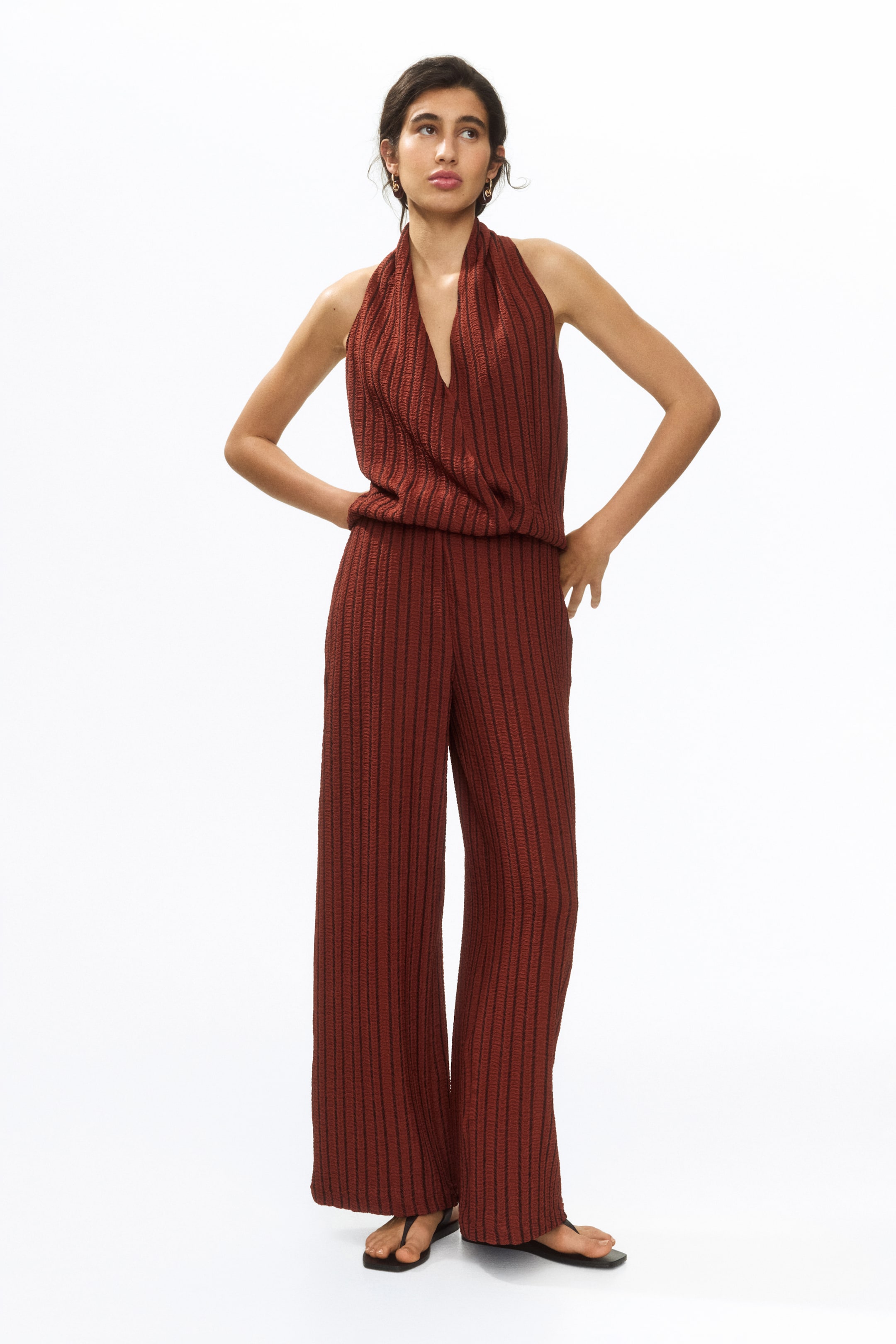 Ampliar la imagen: Un mono color óxido, presentado desde una vista frontal, cuenta con un top de escote halter estilo blusón, pantalones de pierna ancha y un patrón de rayas verticales texturizadas en todo el conjunto.