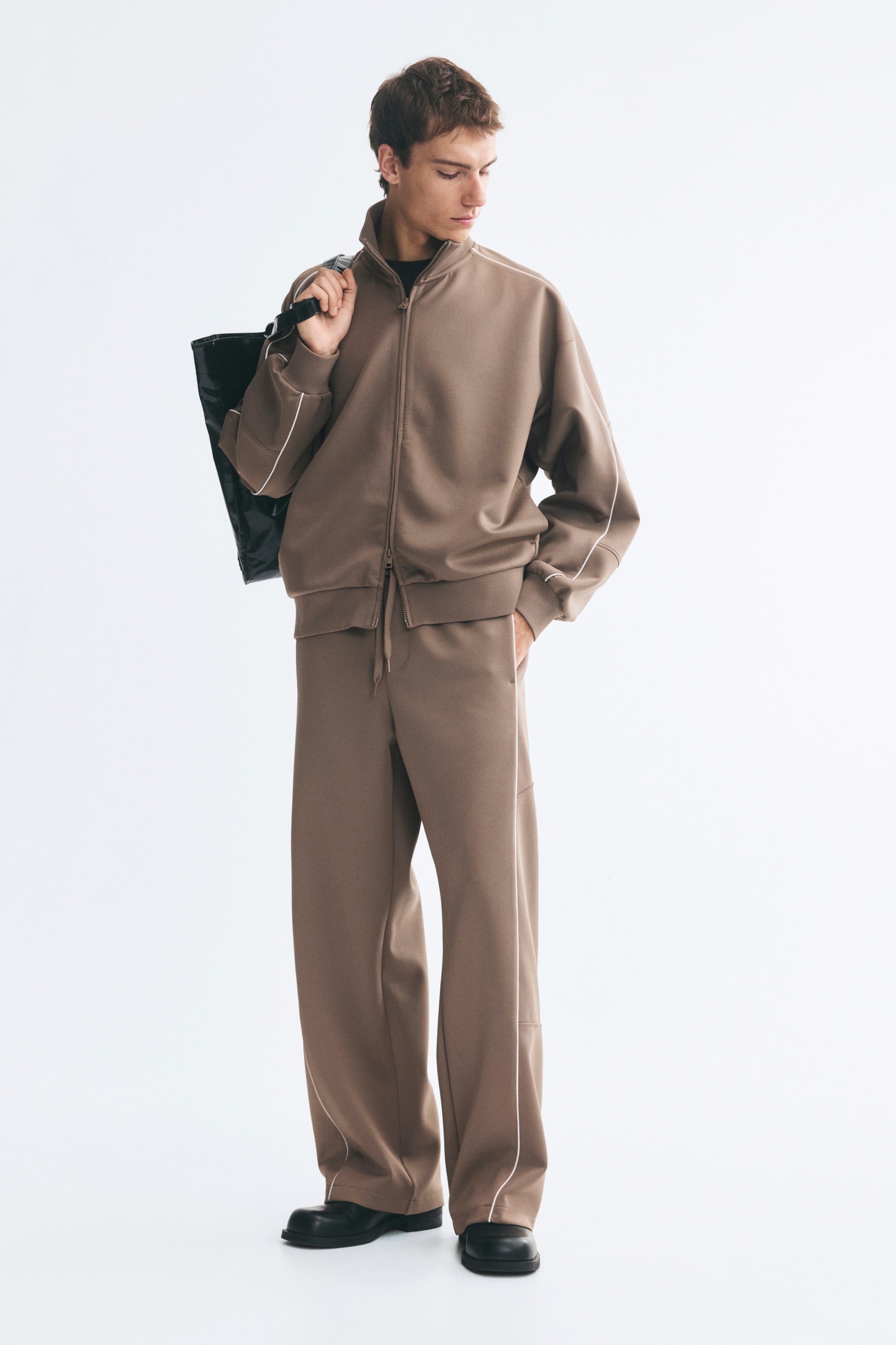 Grotere afbeelding bekijken: Een oversized trainingsjack van bruine scuba-stof met witte contrasterende bies wordt gedragen door het model, gecombineerd met bijpassende wide-leg broek en een zwarte tas over één schouder. Het jack heeft een hoge opstaande kraag, een ritssluiting aan de voorkant en geribde afwerkingen aan de manchetten en zoom.