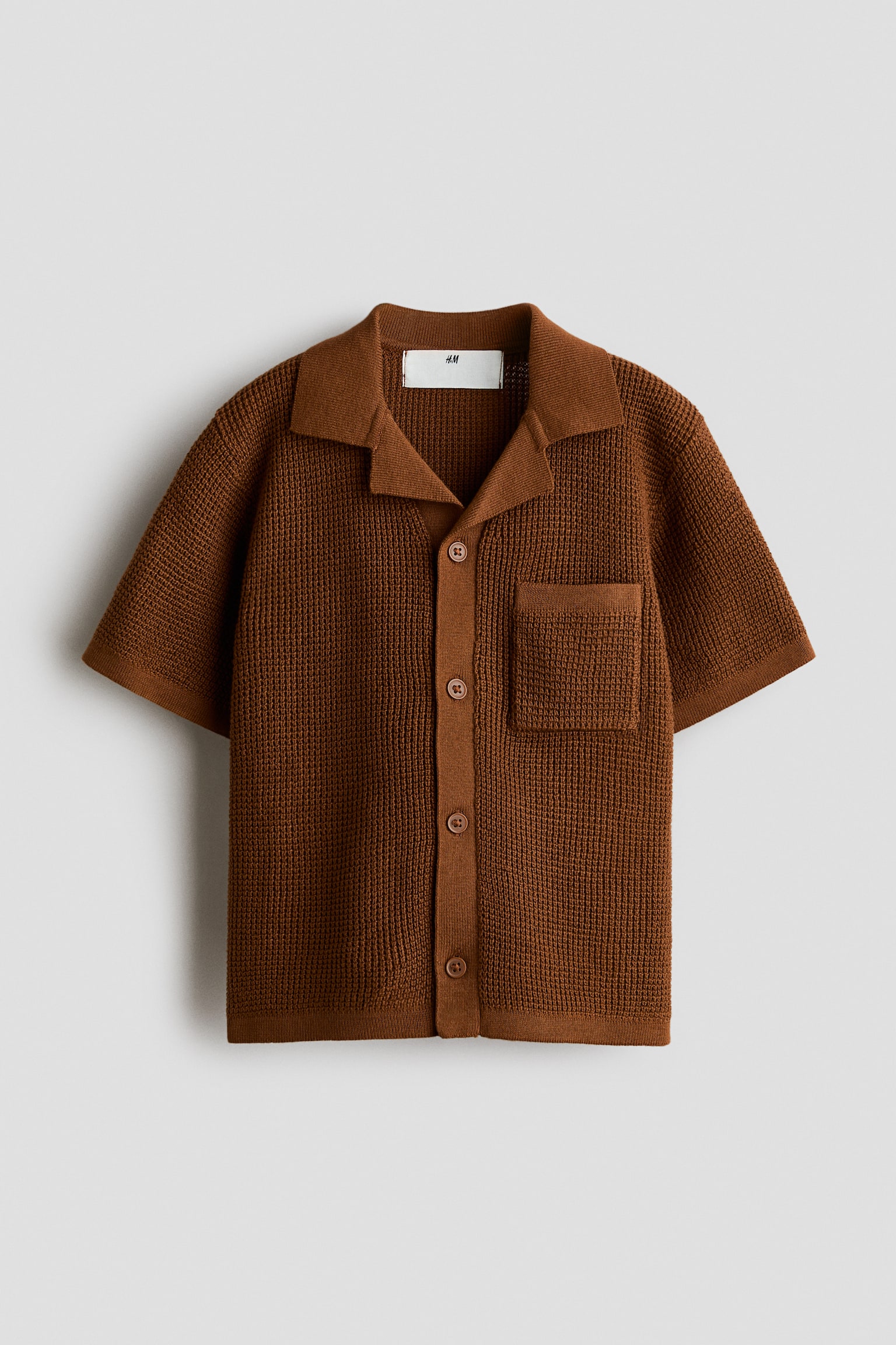 A brown textured knit short-sleeve button-down shirt is shown from the front, featuring a camp collar, button-front fastening, and a single patch pocket on the left chest. The shirt is designed with a waffle-knit structure, ribbed cuffs, and a straight ribbed hem.