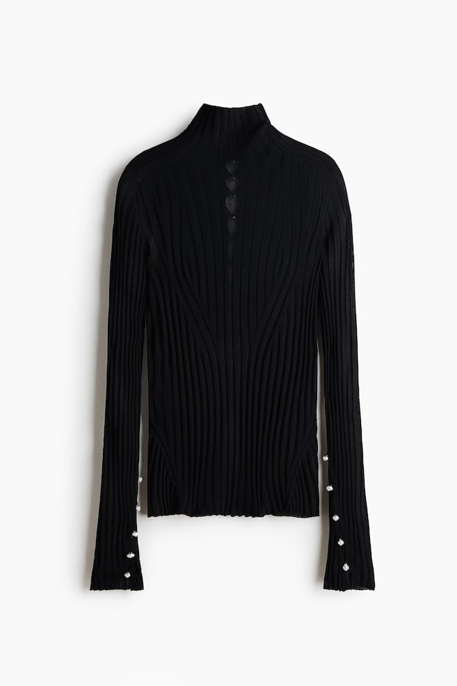 Um top de malha canelada preto exibe uma gola alta com três recortes em forma de coração nas costas. O torso apresenta canelado diagonal, e as mangas compridas têm punhos abertos com cinco pequenos botões de pérola ao longo de cada lado. - 1