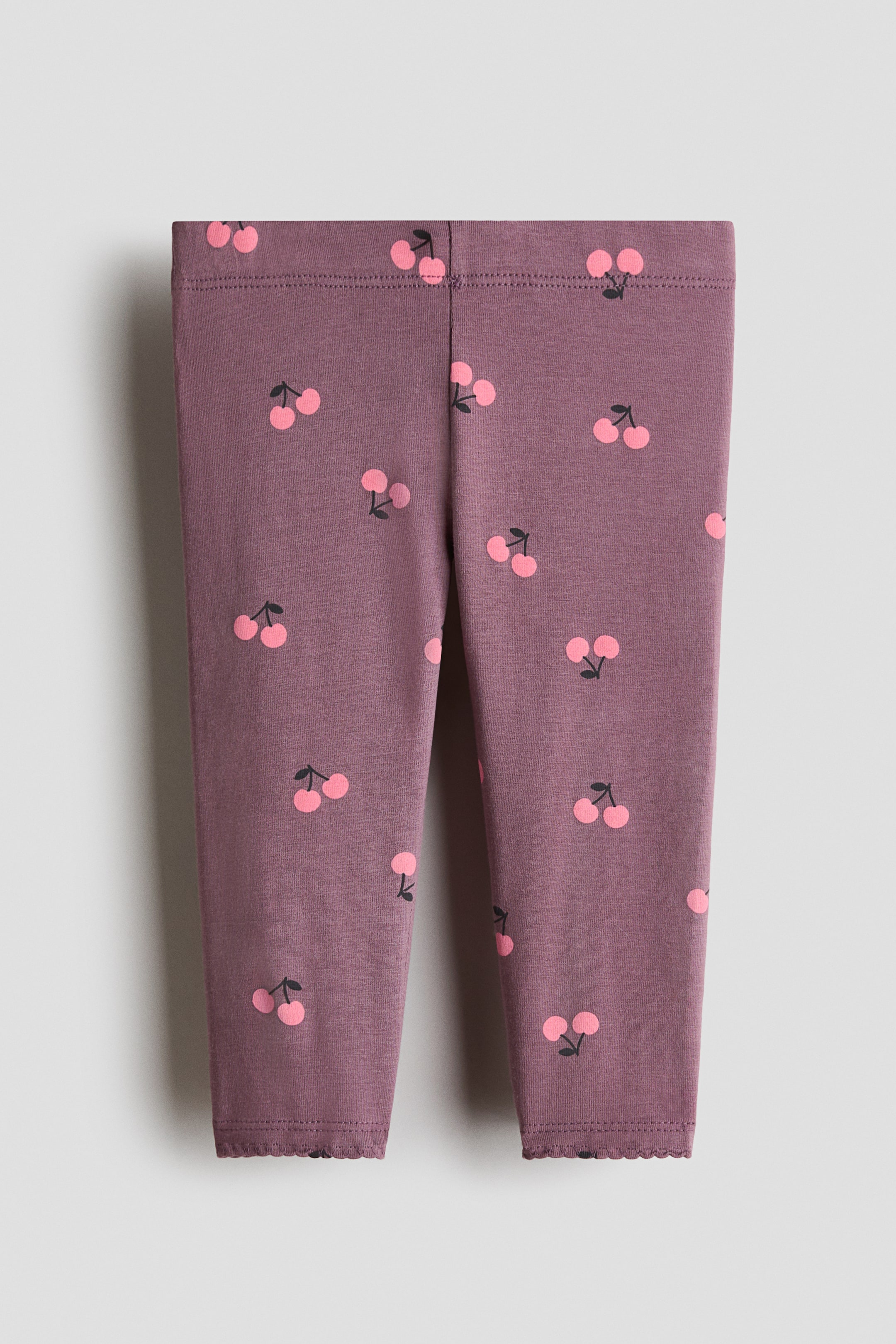 View larger image: Leggings are exhibiting a front-facing perspective, showcasing a printed jersey fabric adorned with pink cherries on a mauve backdrop. They feature an elasticated waistband and scalloped hems.