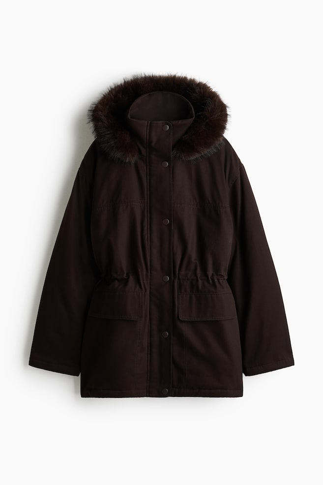 Vue de face d'une parka oversize marron foncÃ©, dotÃ©e d'une capuche ornÃ©e d'une bordure en fausse fourrure moelleuse. La veste est conÃ§ue avec un rabat coupe-vent Ã  boutons-pression recouvrant une fermeture zippÃ©e frontale, une taille froncÃ©e Ã  cordon de serrage et deux grandes poches plaquÃ©es Ã  rabat sur l'avant. - 1