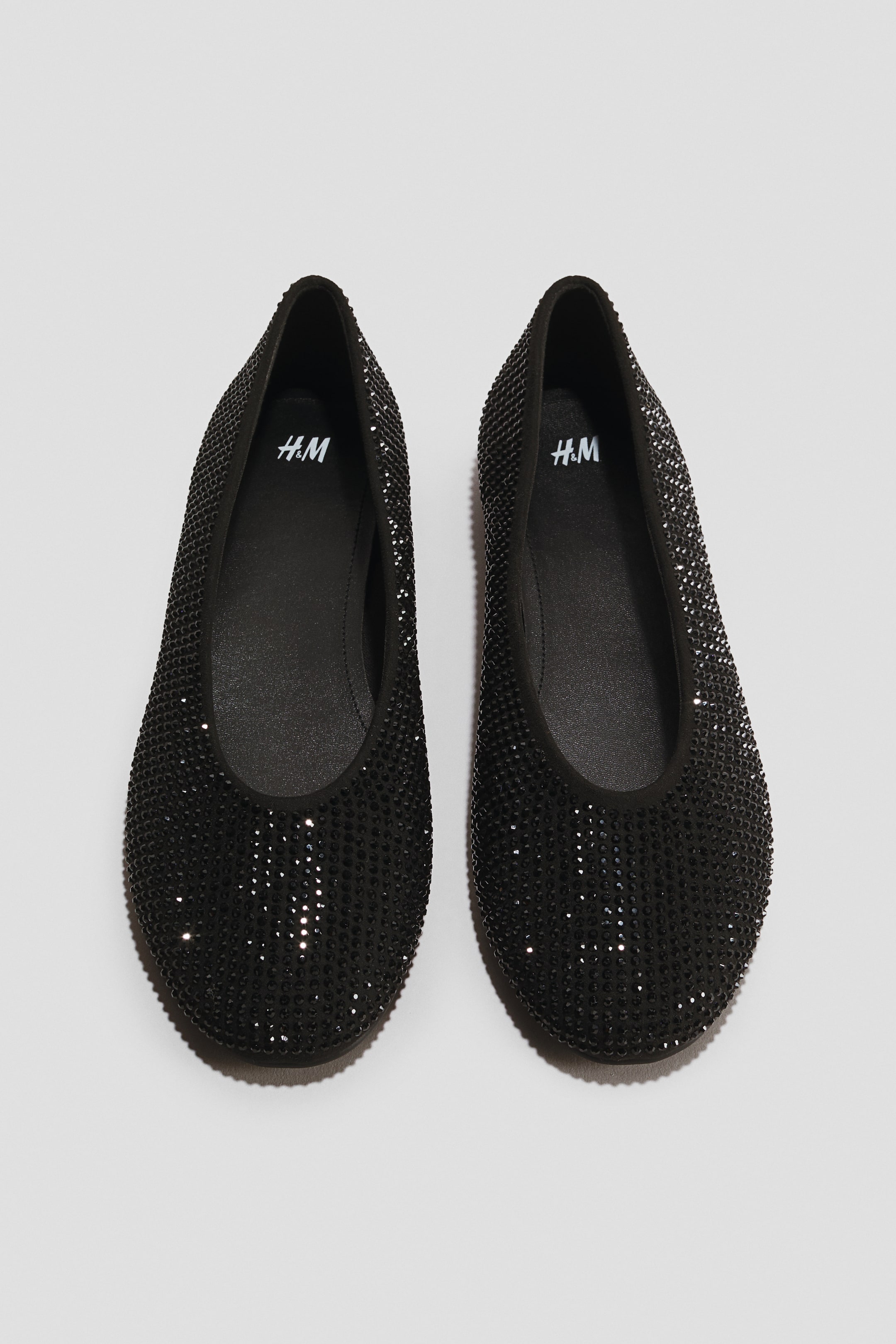 Ver imagen más grande: Un par de bailarinas negras aparece orientado hacia el frente en una vista cenital, con el corte completamente adornado con pequeños brillantes negros. Los zapatos presentan un diseño de punta redondeada, ribete negro de tejido alrededor del cuello y plantillas negras impresas con un logotipo blanco.