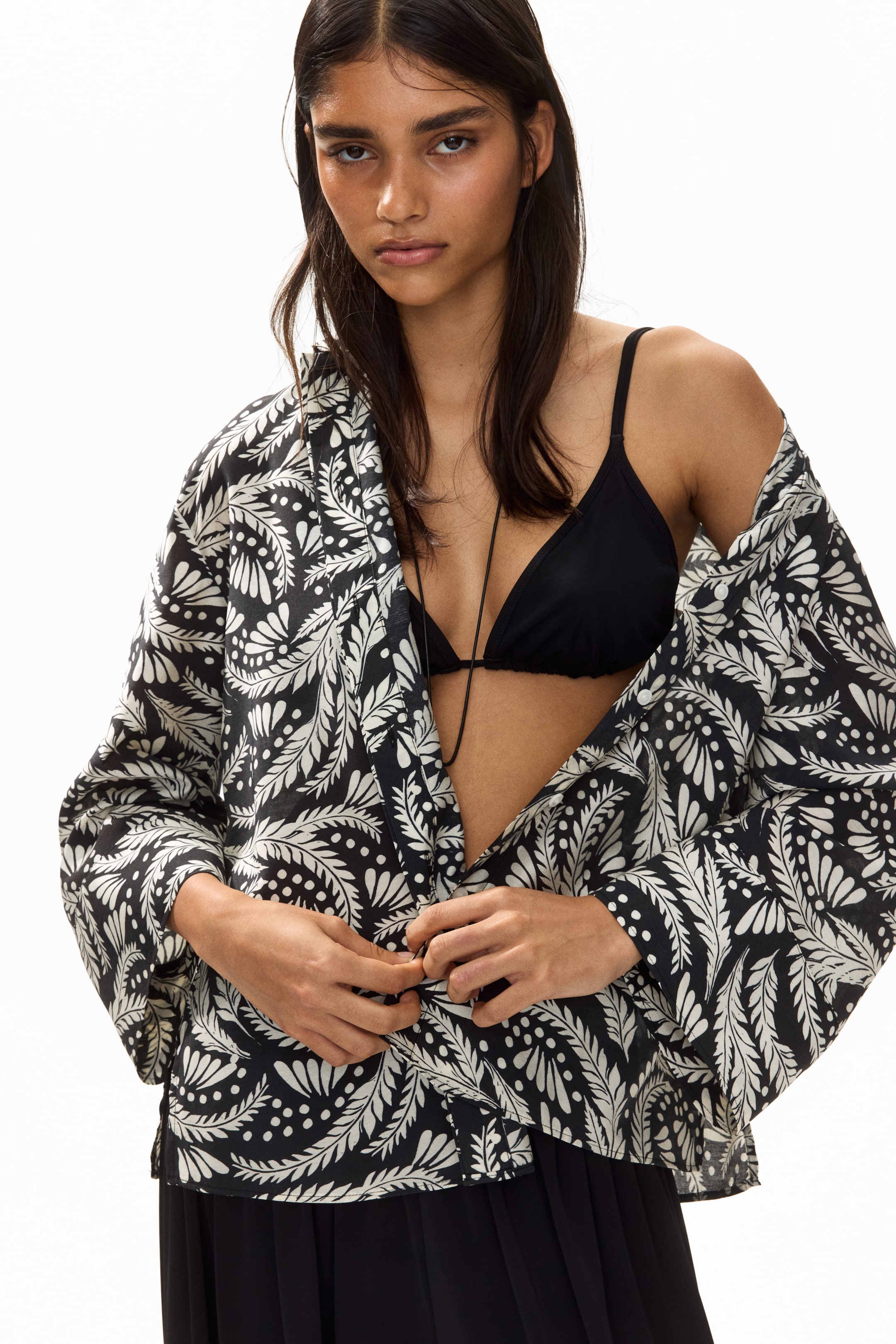 View larger image: The model is showcasing a shirt crafted from a viscose-blend weave adorned with a black and white botanical print. This piece features a collar, dropped shoulders, long sleeves with wide cuffs, and a straight-cut hem. The model is seen adjusting the concealed front buttons of the shirt, which is styled open over a black bra.