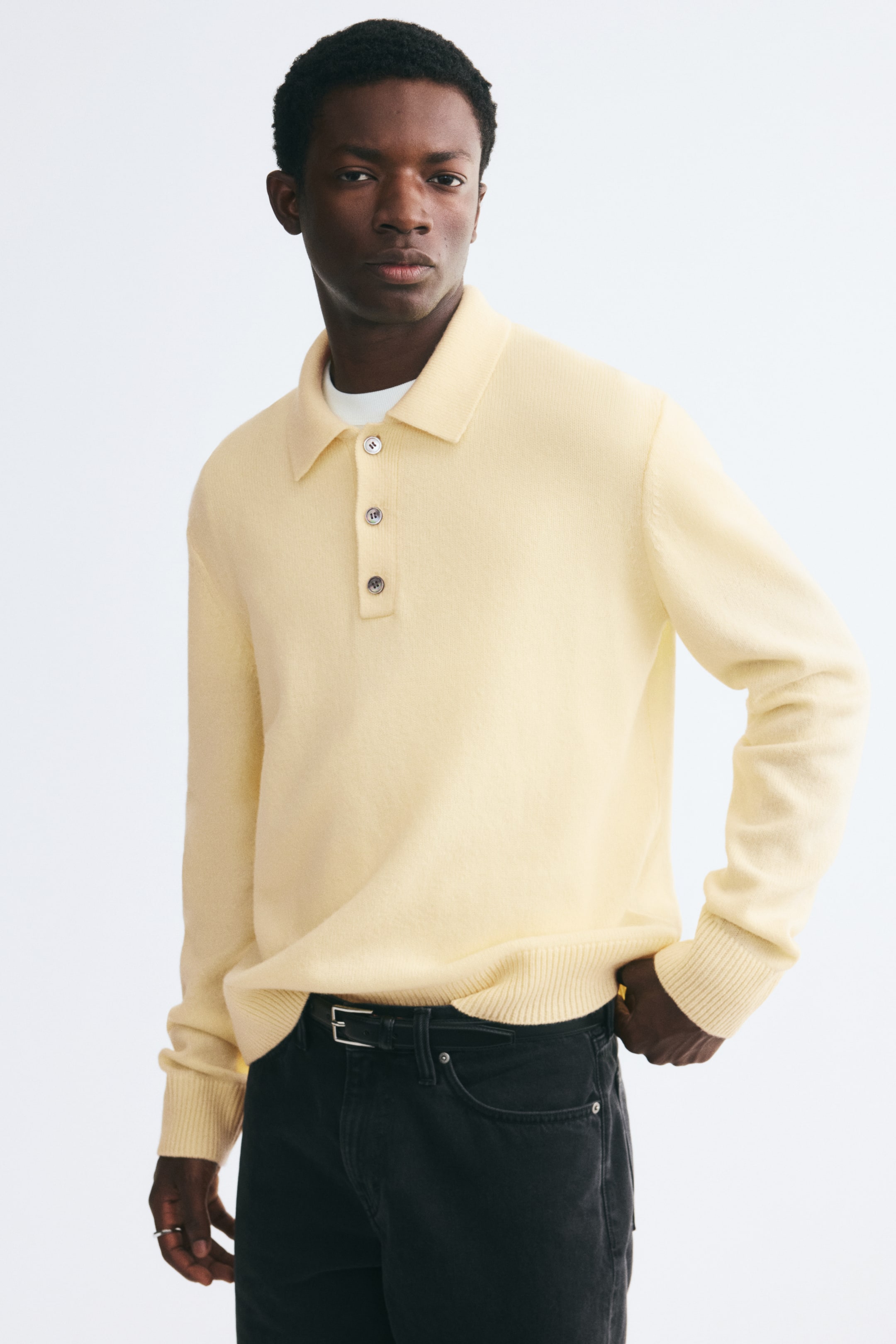 View larger image: The model is wearing a light yellow, fine-knit, long-sleeve polo shirt layered over a white T-shirt and tucked into dark jeans. The shirt features a classic collar, a three-button placket, and ribbed detailing at the cuffs and hem.