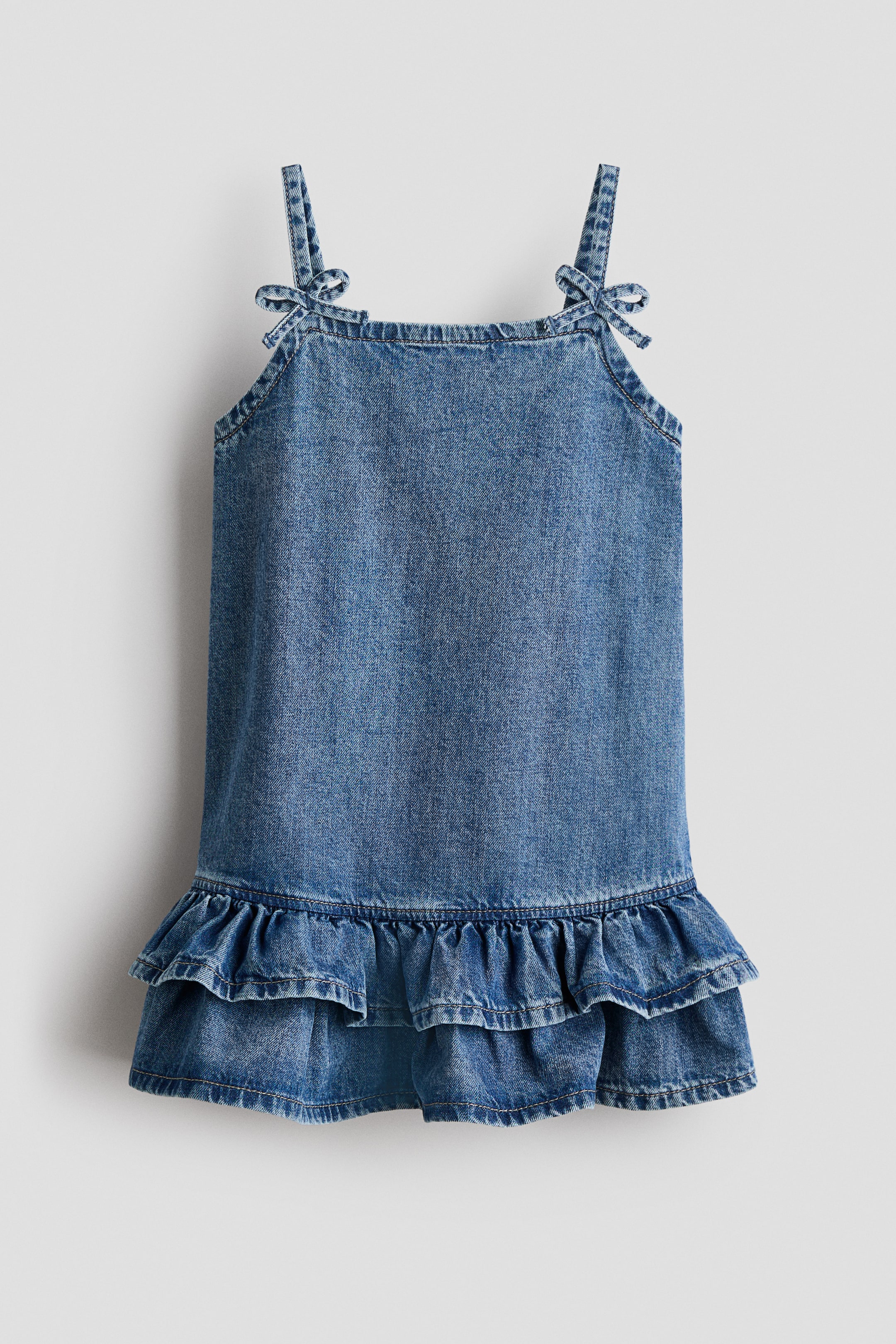 View larger image: Denim dress, FRONT, features a blue wash with tie-up shoulder straps. It showcases a straight body and two tiers of ruffles at the bottom hem.