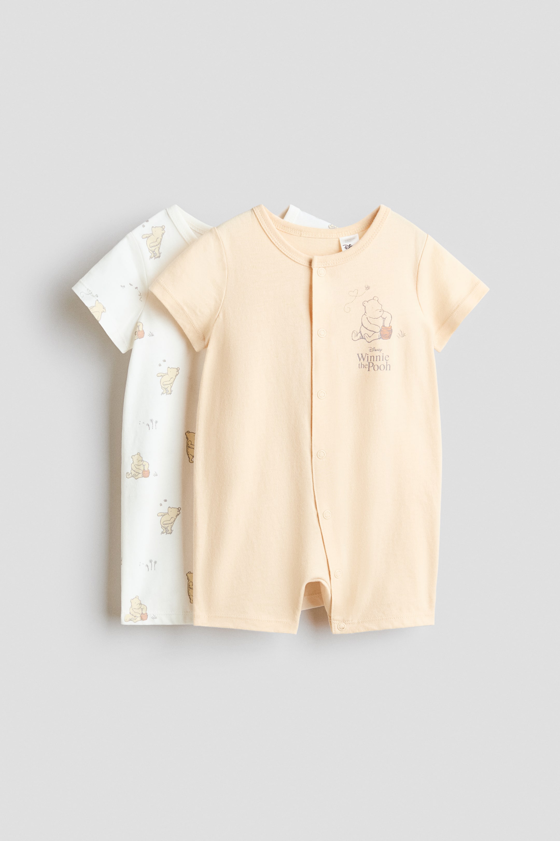 View larger image: Two baby rompers are on display: a front-facing light orange romper with short sleeves, a round neckline, and a full snap button placket running from the center front to the inner leg, featuring a Winnie the Pooh graphic and text on the chest. Behind it, a partially visible off-white romper showcases an all-over pattern of Winnie the Pooh characters.